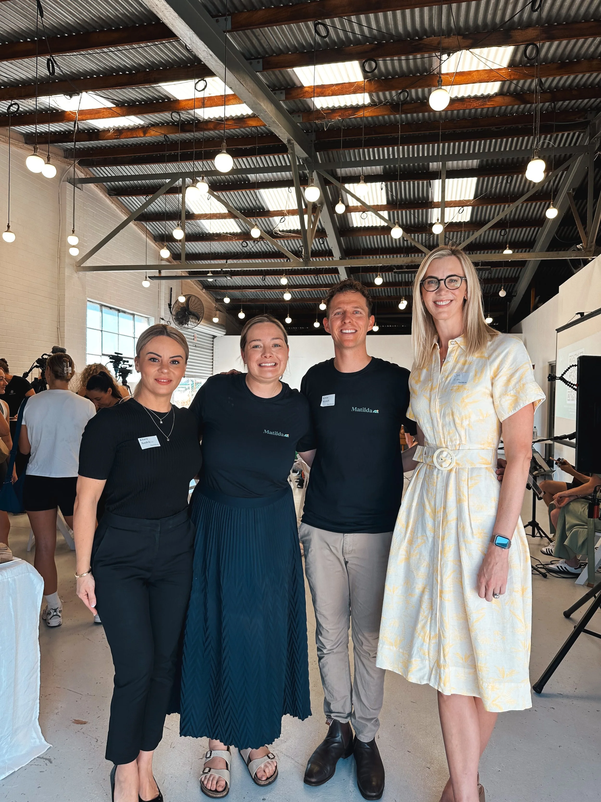 Four smiling people standing together in a large industrial-style room with hanging lights, a corrugated metal ceiling, and visible crowd in the background. Three women and one man, all wearing casual or semi-formal attire.