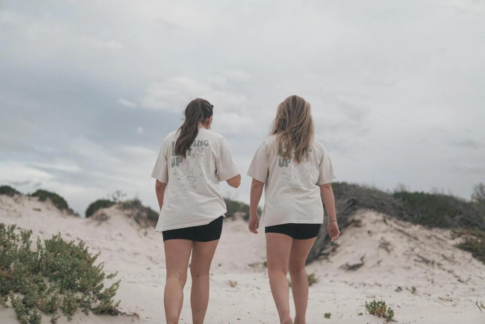 Two women walking away on a sandy beach with dunes and sparse vegetation, under a cloudy sky.