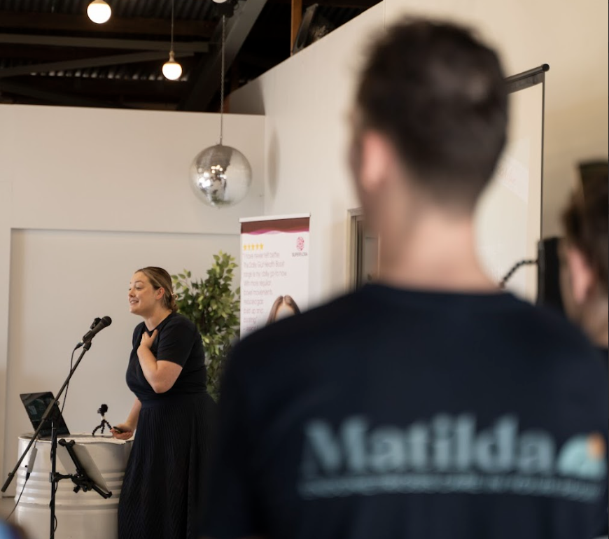 A woman is speaking at a microphone in front of an audience, smiling and gesturing with her hand on her chest. There are two people in the foreground, one with the back of their head visible, wearing a shirt with the word 'Matilda' on it. The background includes a white wall, a potted plant, a banner, and a shiny disco ball hanging from the ceiling.