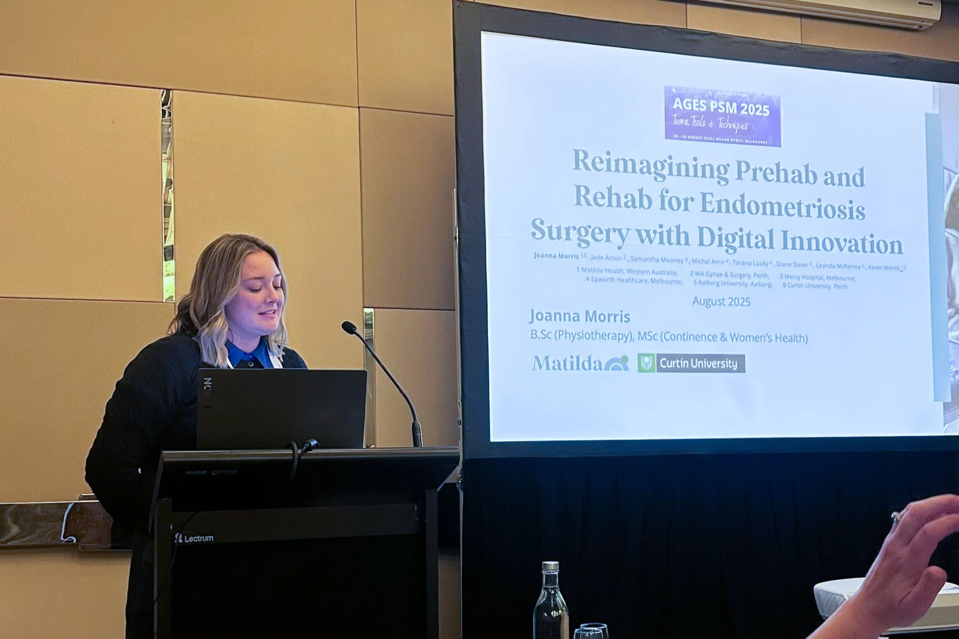 A woman with blonde hair, wearing a blue shirt and black blazer, standing at a podium with a laptop, speaking into a microphone during a conference. Behind her is a large screen displaying a presentation slide titled 'Reimagining Prehab and Rehab for Endometriosis Surgery with Digital Innovation' with authors and institutional affiliations.