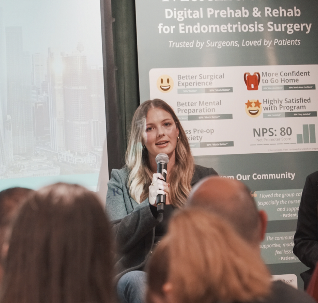 Young woman speaking into a microphone during a presentation on endometriosis surgery and rehabilitation, with a large poster displaying statistics and positive outcomes in the background.