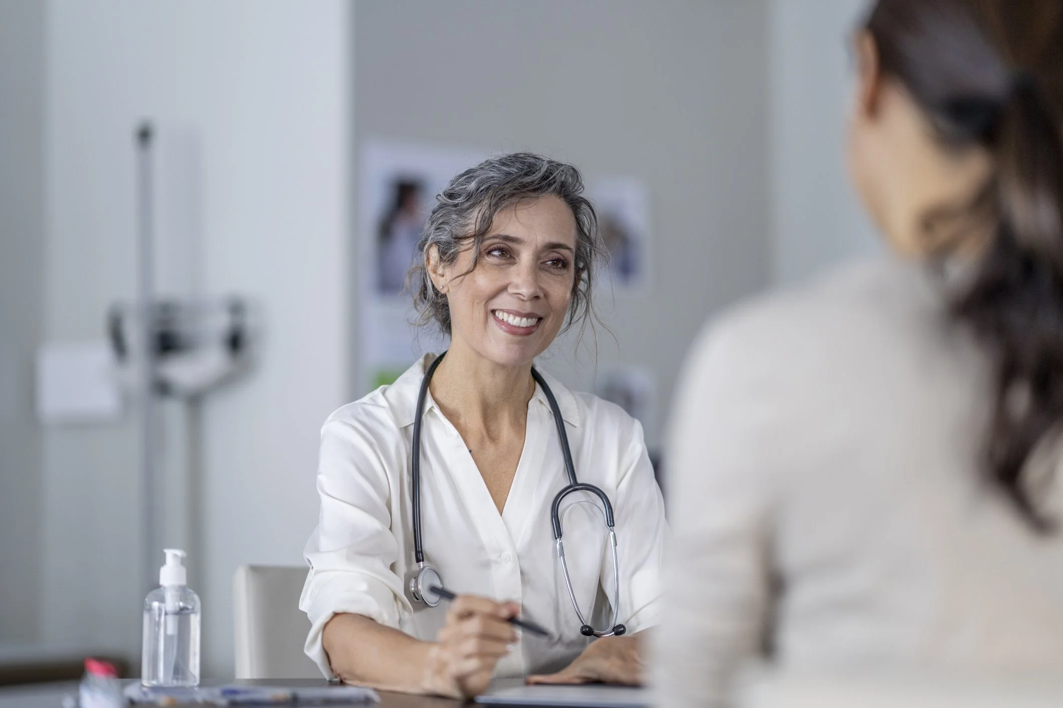 A female doctor with gray hair and a stethoscope around her neck smiling and talking with a female patient in a medical office.