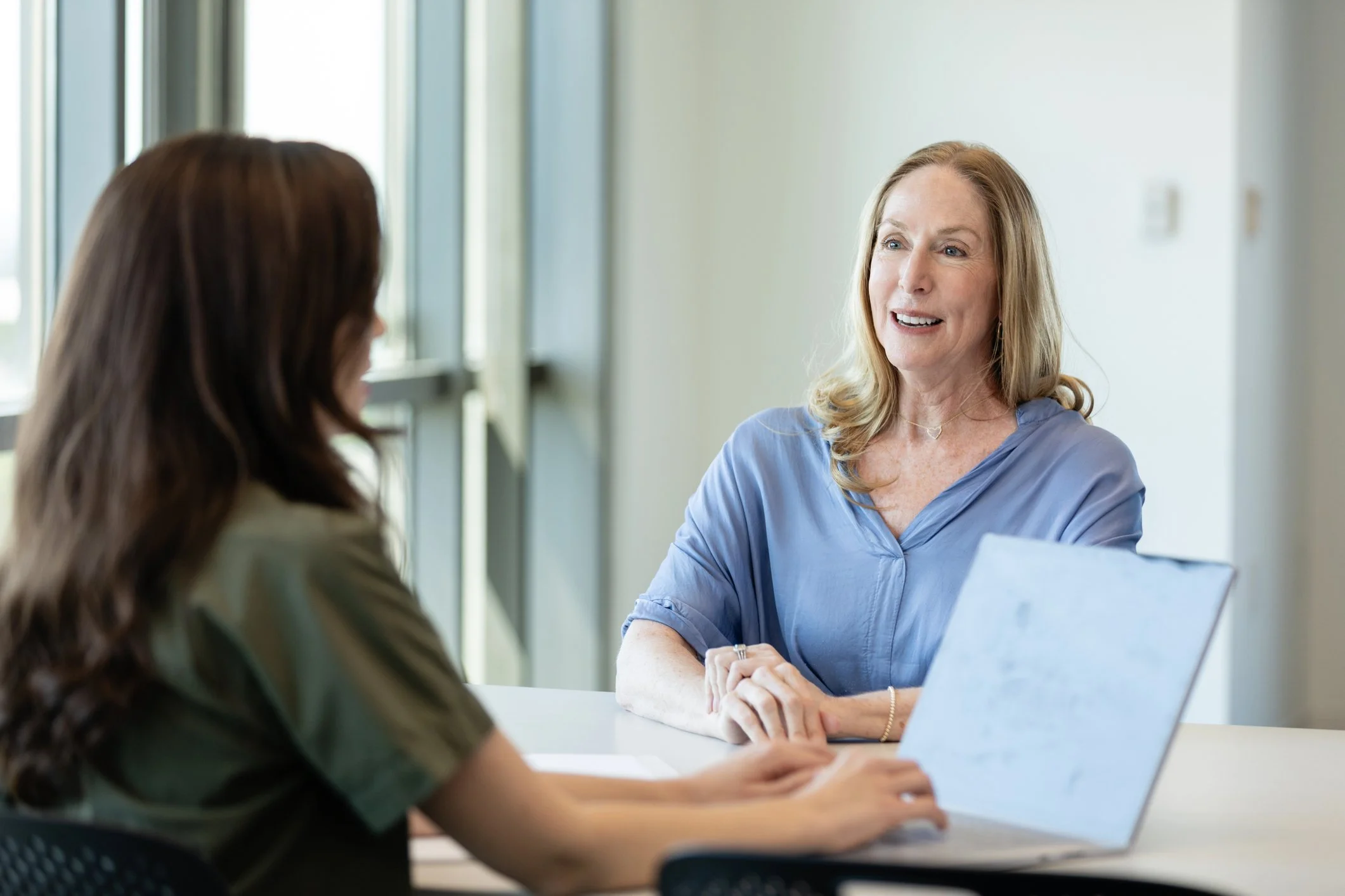 A woman with blonde hair wearing a blue shirt talking to a woman with brown hair in an office setting.
