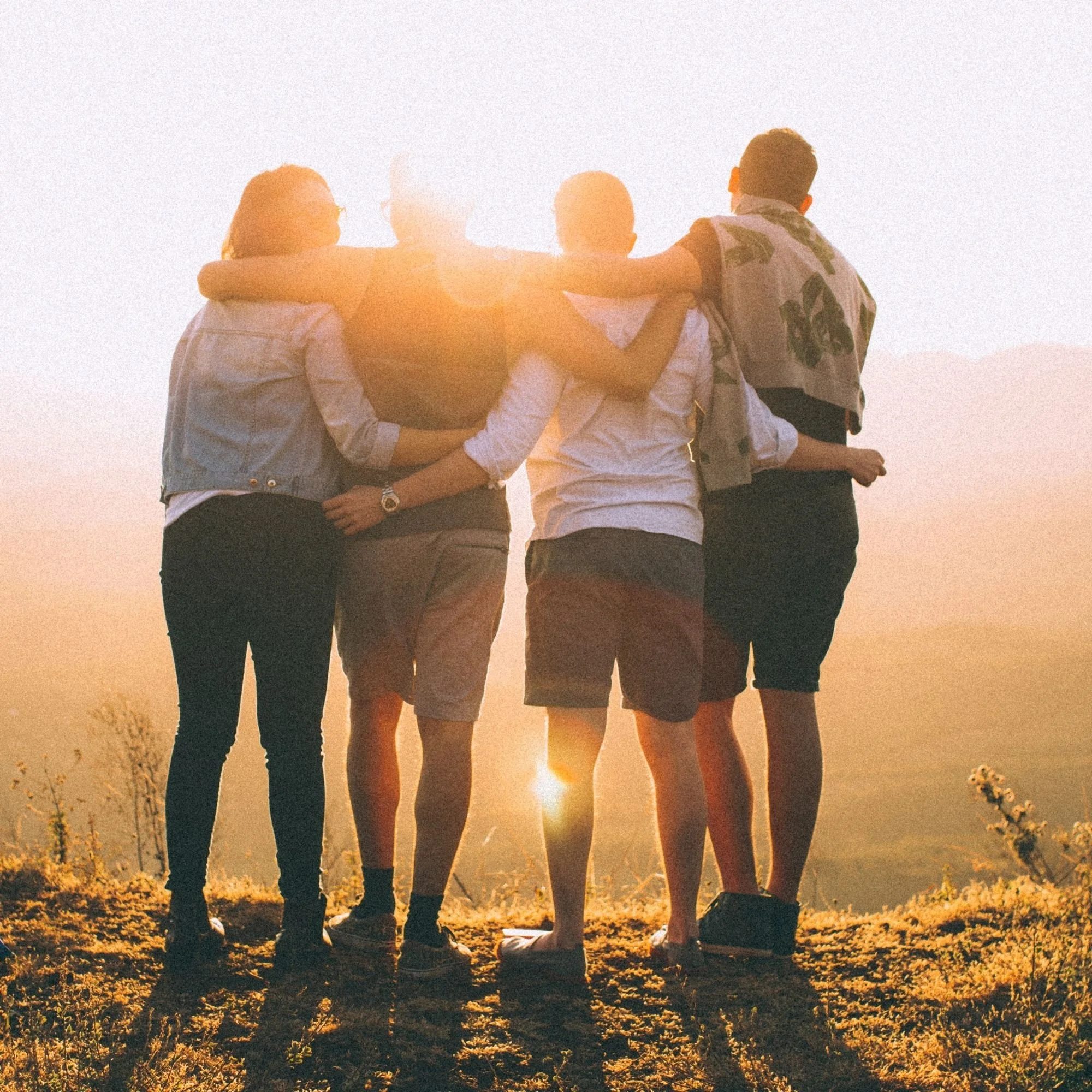 Four friends sitting outdoors at sunset, with their arms around each other, enjoying a peaceful moment.