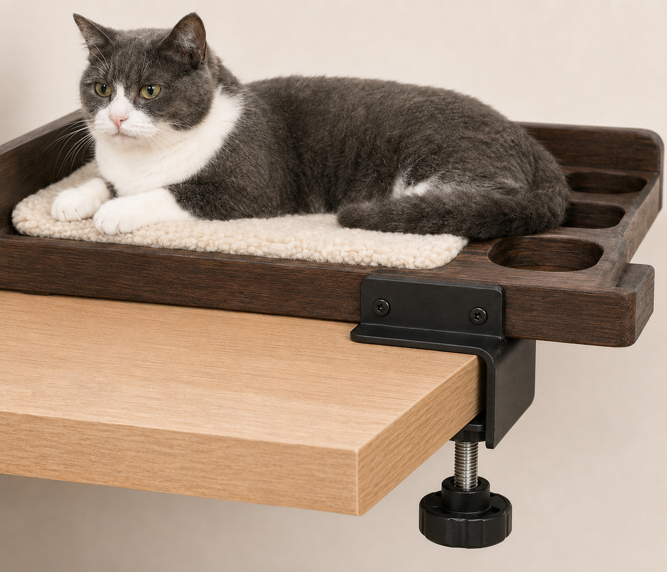 A gray and white cat laying on a beige fleece mat inside a dark wooden cat perch mounted on a light wooden table.