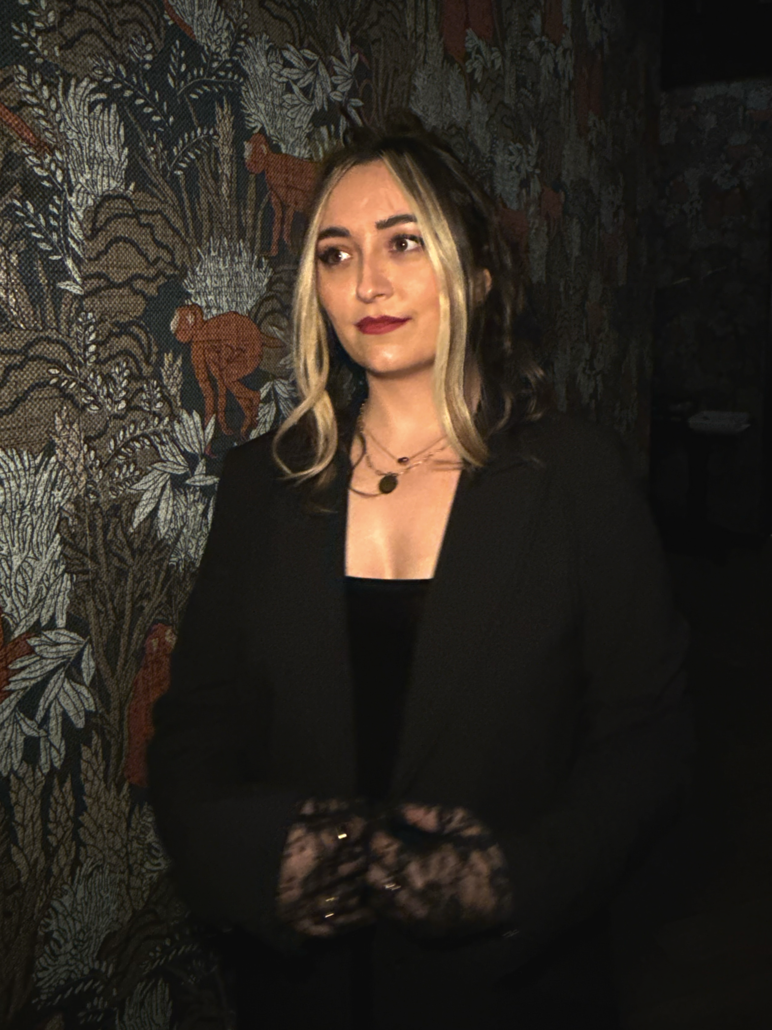 A woman with blonde and dark hair wearing a black outfit, necklace, and lace shoes, standing indoors against a patterned tapestry or wall hanging.
