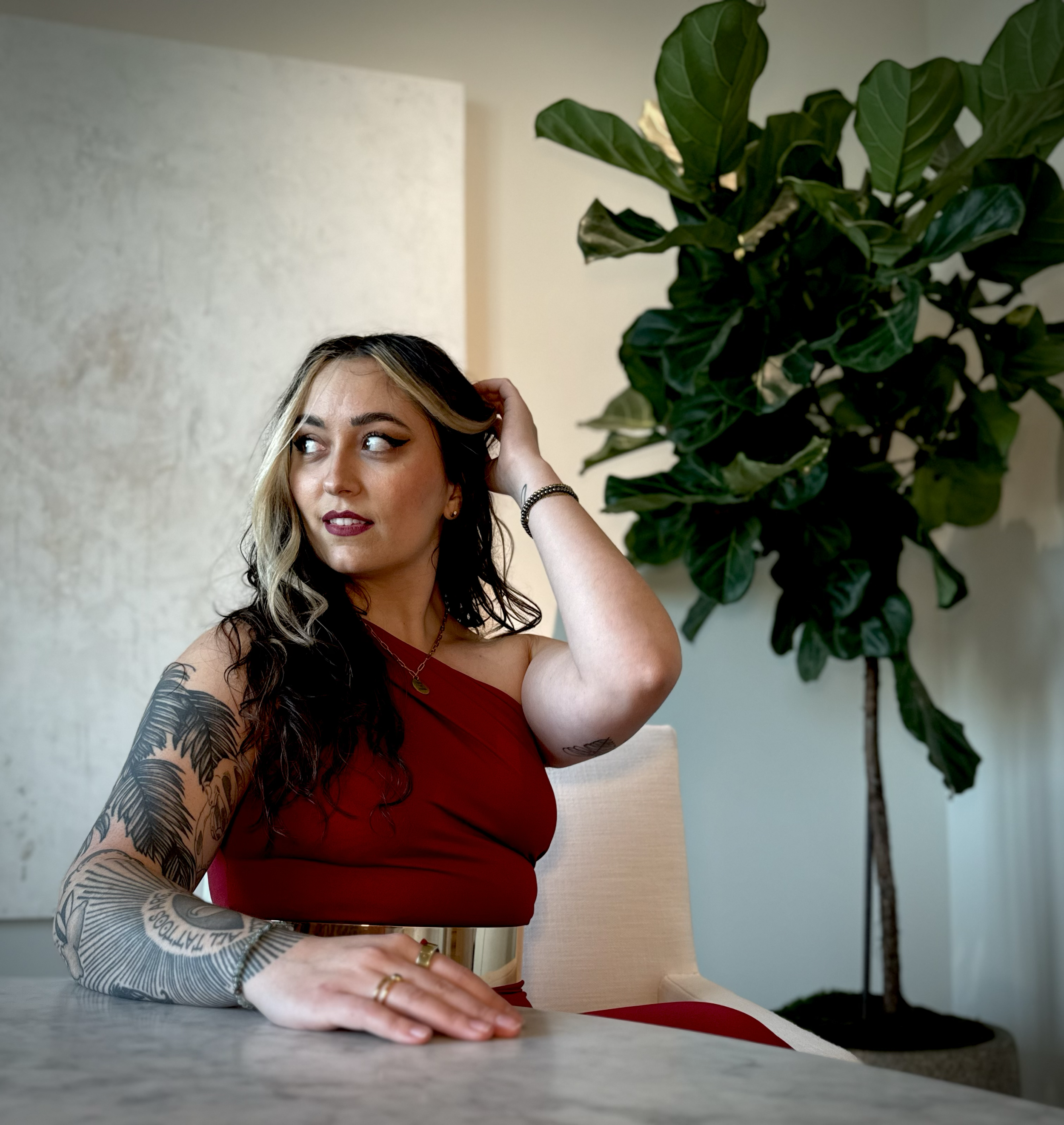 A woman with tattoos on her left arm wearing a red dress sitting at a table with her left hand on the surface, and her right hand adjusting her hair. She has dark, wavy hair with blonde highlights and is wearing makeup. Behind her is a large green leafy plant, a table lamp, and a white textured wall.
