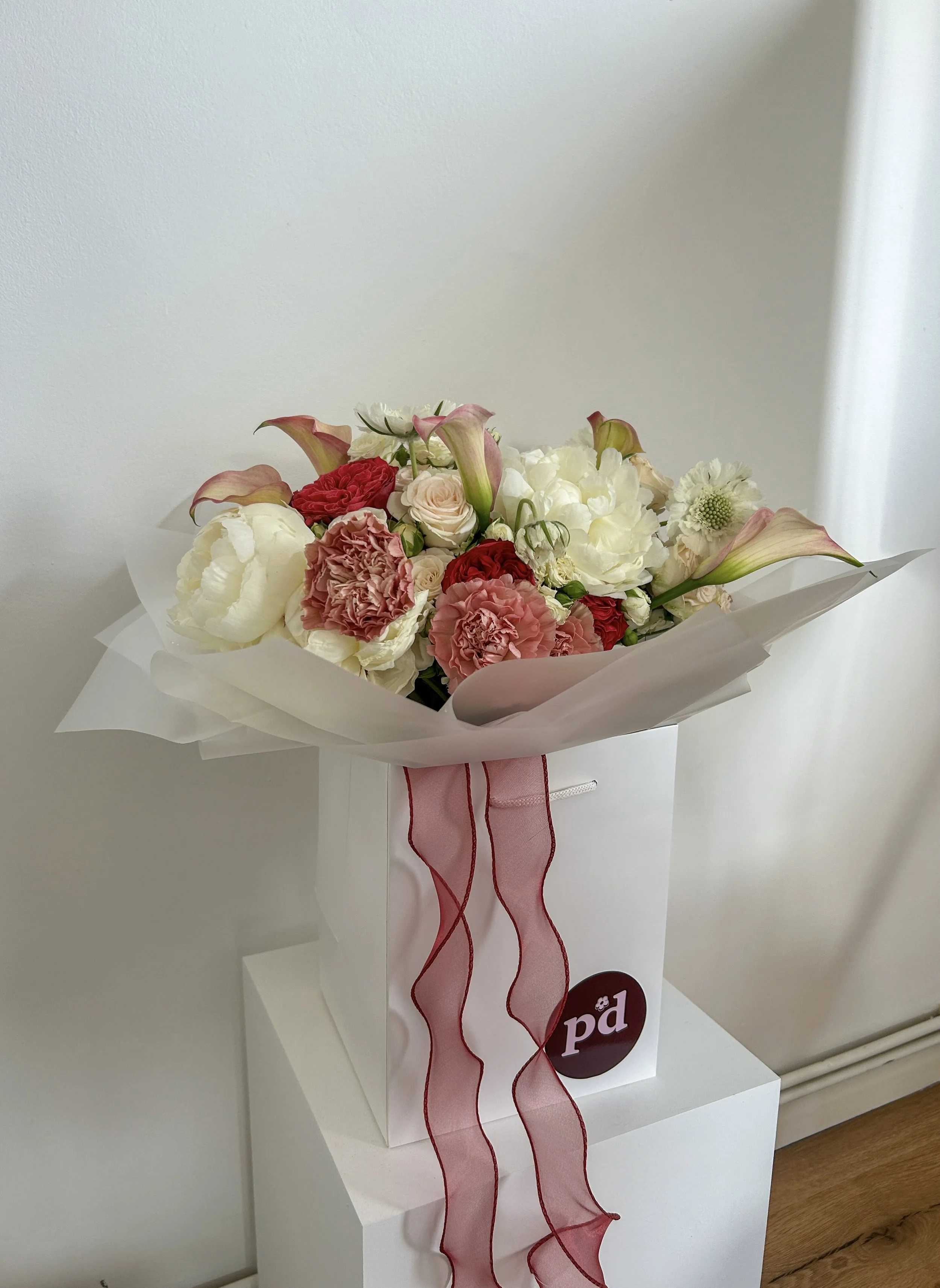 A bouquet of various flowers, including white, pink, and red roses, along with pink calla lilies and other blooms, wrapped in white paper with a pink and red ribbon, displayed on a white stand, made by Petal'd