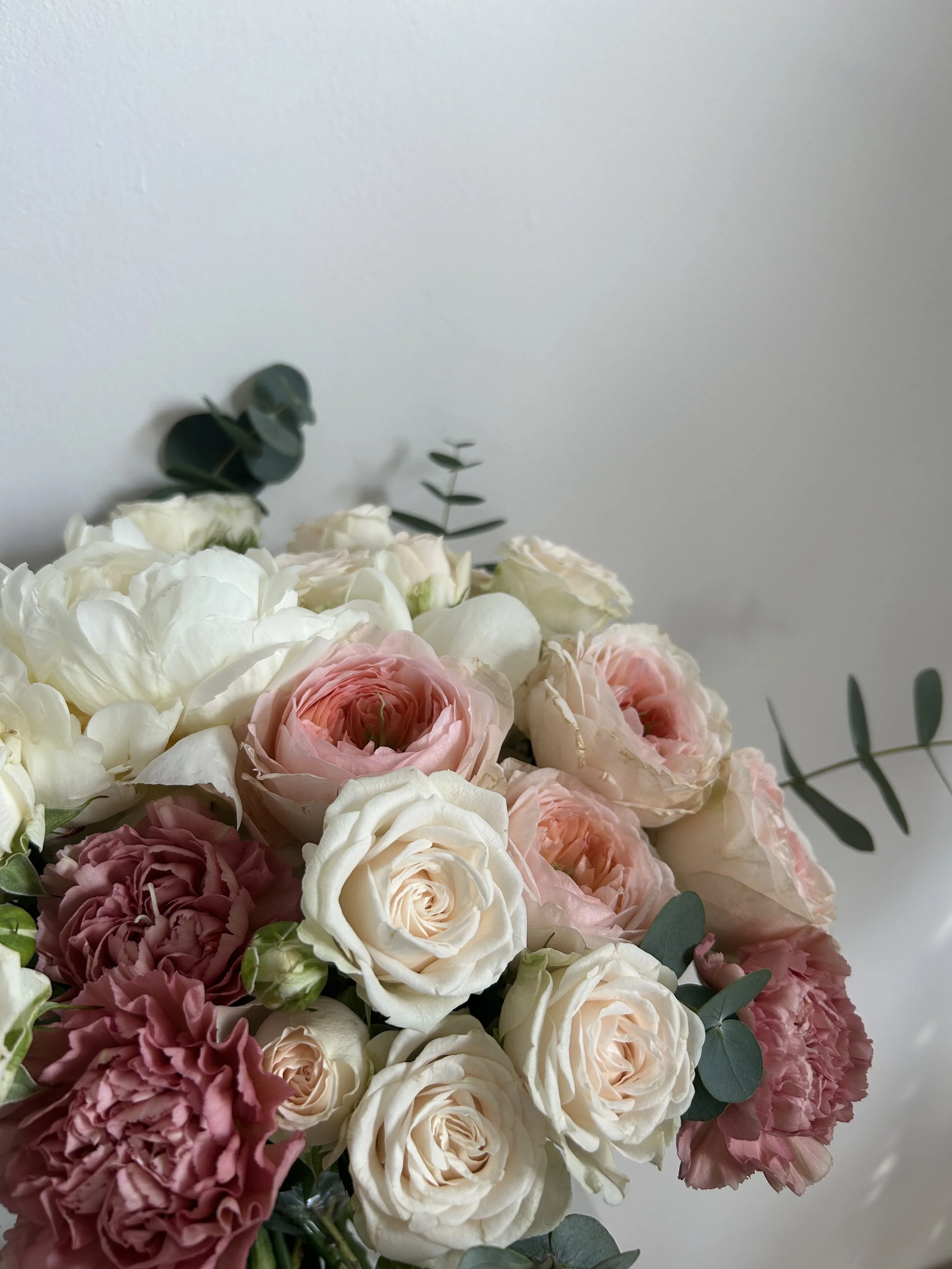 A bouquet of various flowers including white roses, pink roses, and pink carnations, with eucalyptus leaves against a plain gray wall background. Made by Petal'd