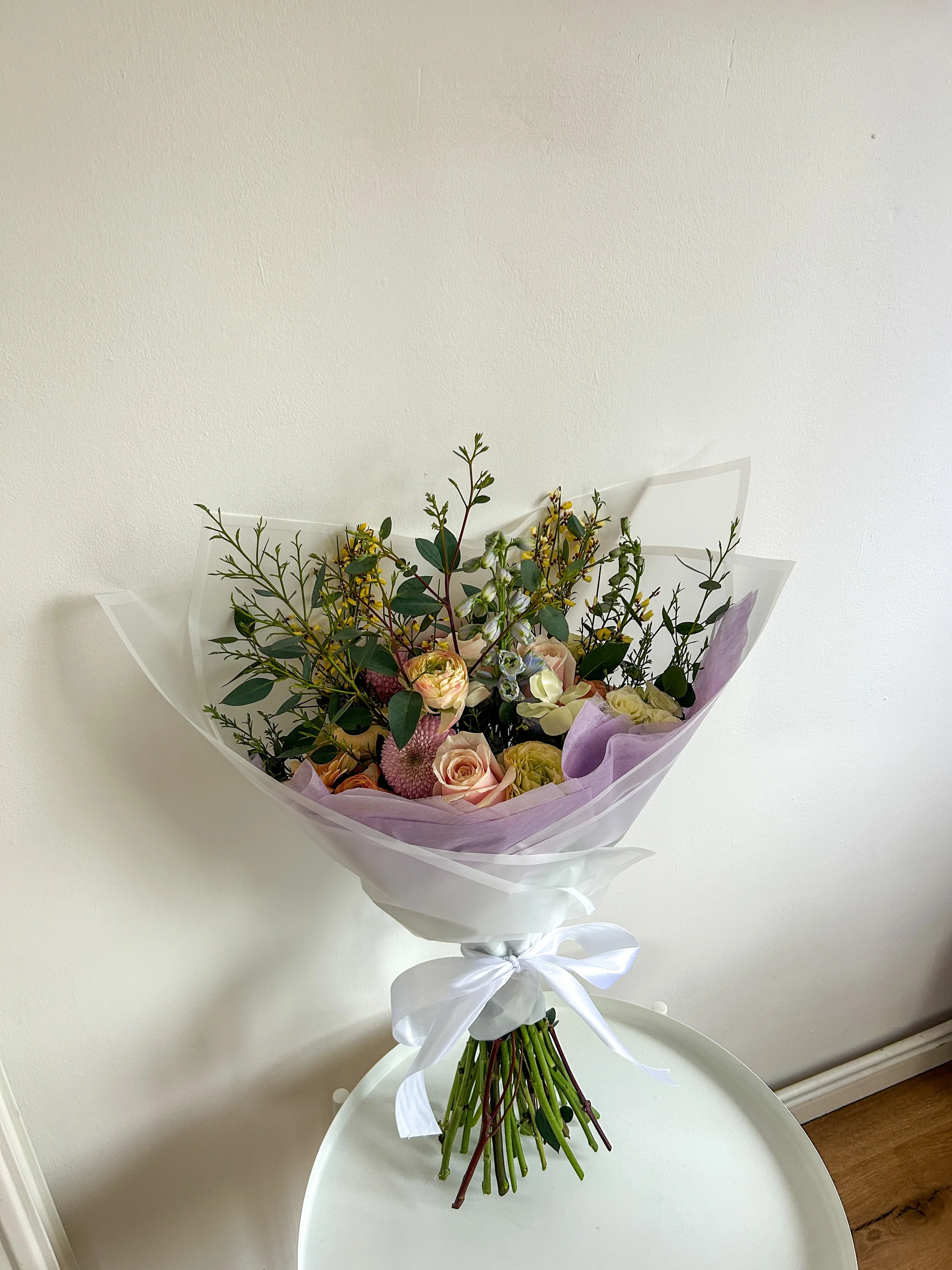 A bouquet of various flowers, including roses, in pastel shades, wrapped in purple and clear paper, tied with a white satin ribbon, sitting on a white round table against a plain white wall.