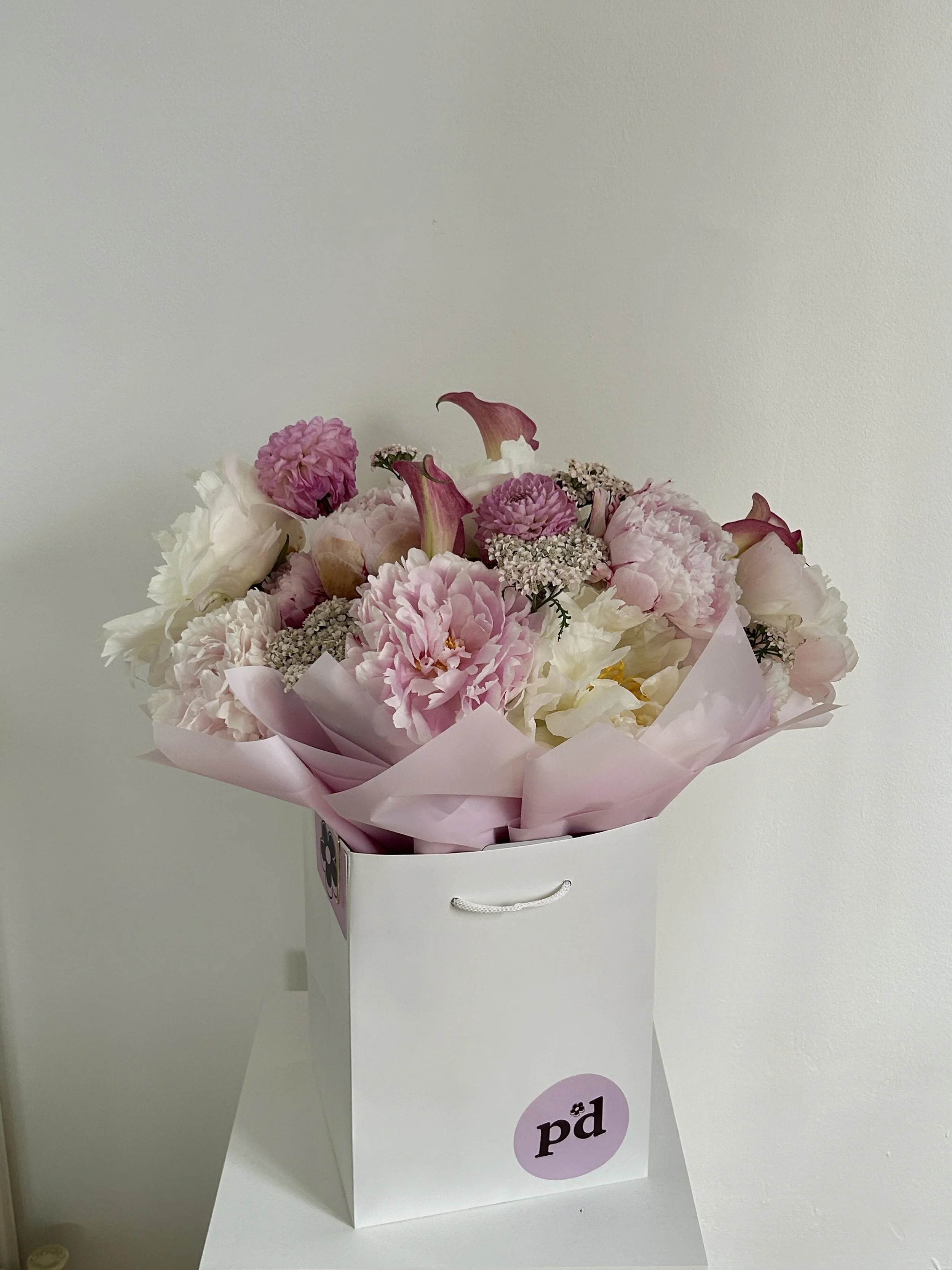 A white gift bag with black and purple 'pd' logo logo, filled with pink and white flowers, on a white surface against a plain wall.