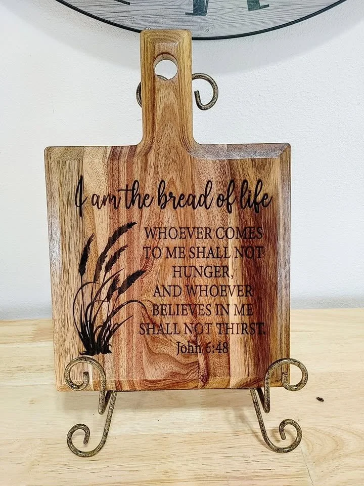 A wooden decorative cutting board with a religious quote from John 6:48, mounted on a wrought iron stand.