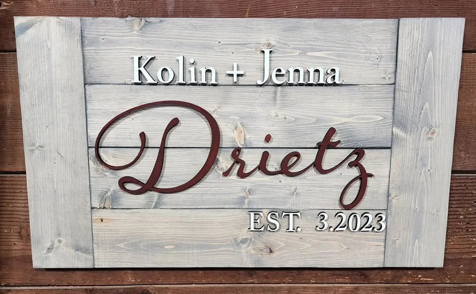 Wooden sign with whitewashed background displaying the names 'Kolin + Jenna Drietz' and 'EST. 3.2023' in metal letters.