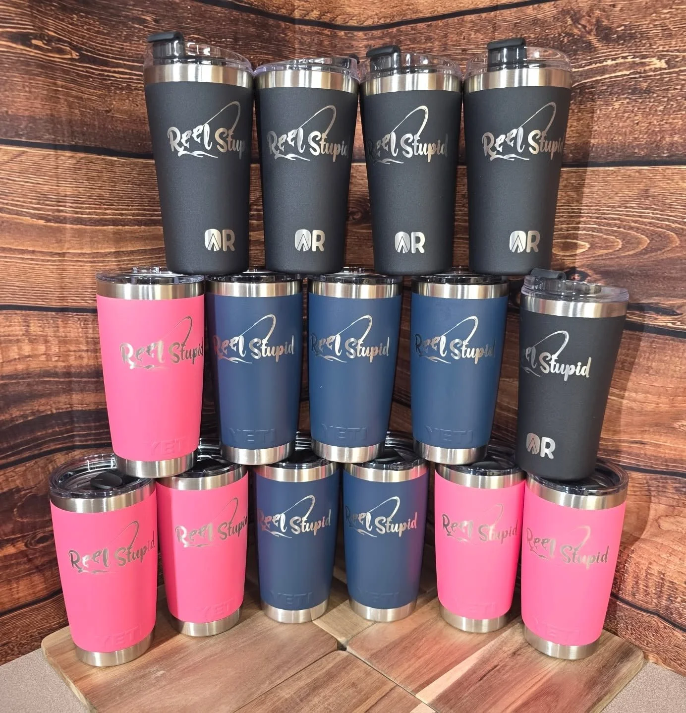 Stacked travel mugs with black, pink, and blue colors, displaying the phrases "Reel Stupid" and a fishing hook logo, set against a wooden background.