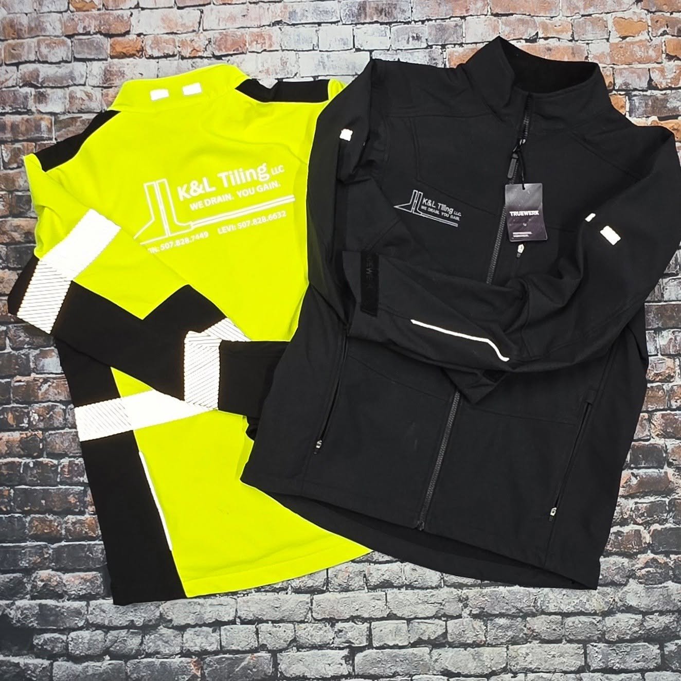A high-visibility yellow safety jacket with black and white reflective stripes and a black zip-up jacket with branding, all laid out on a brick wall background.