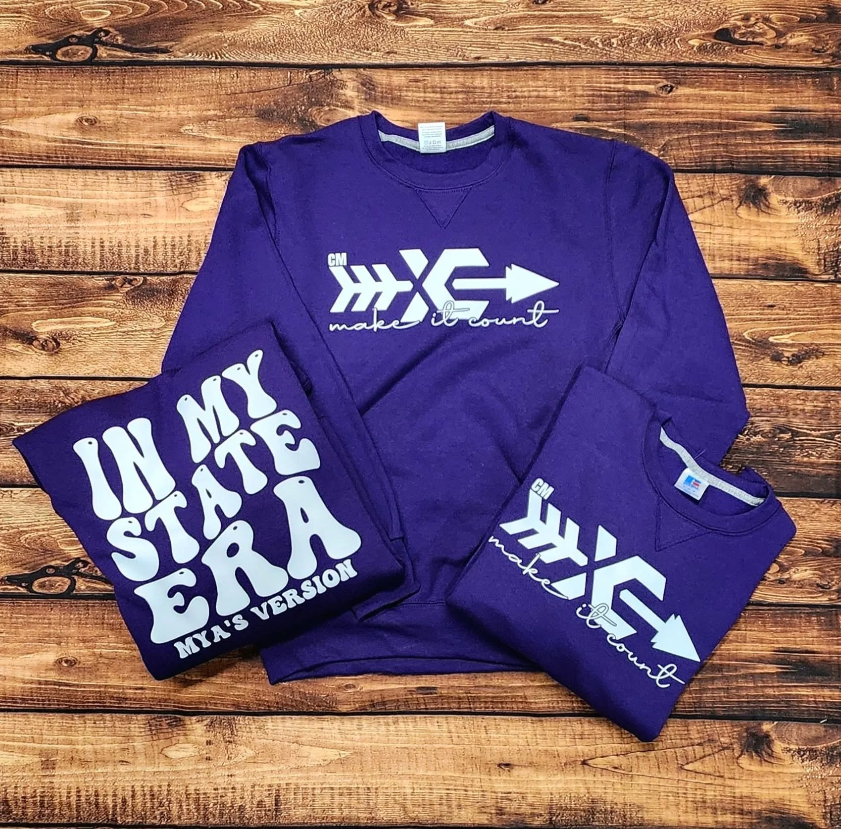A purple sweatshirt with white text on the front that reads 'make it count' accompanied by a white arrow graphic. Next to it, a purple T-shirt with the phrase 'IN THIS STATE' in large white letters. The items are laid out on a wooden surface.