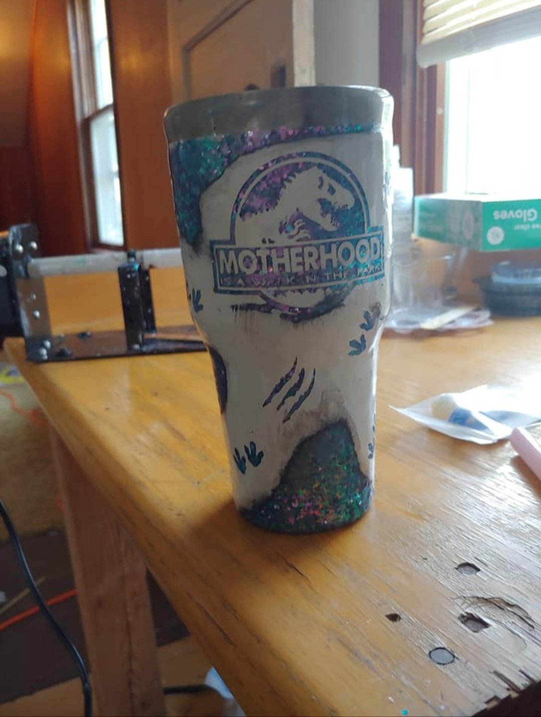 A ceramic mug with the word "Motherhood" and a mountain logo, decorated with colorful holographic accents, placed on a wooden table.