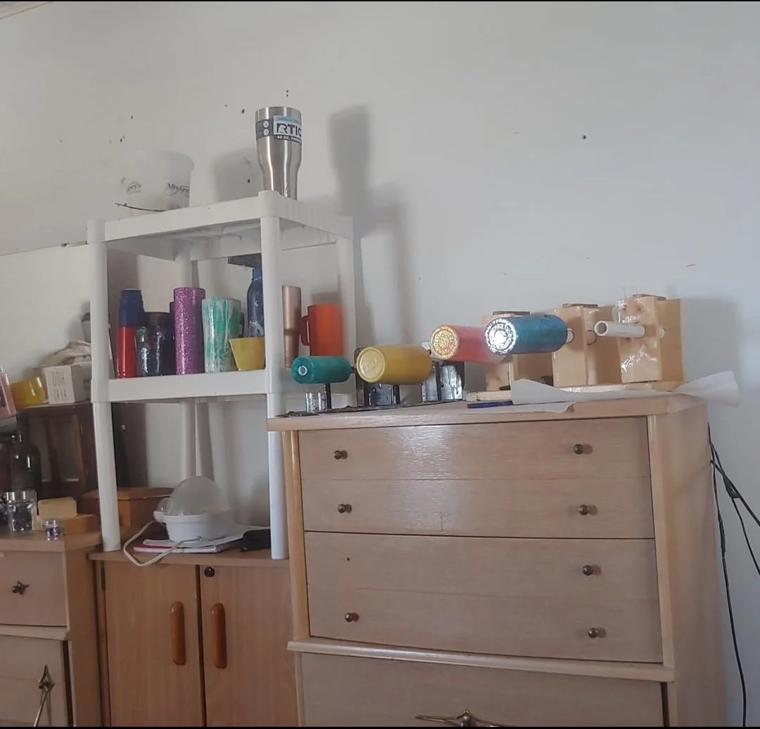 A wooden dresser with several drawers next to a white shelving unit. The dresser's top surface holds several craft foam tubes, some wrapped in paper. The shelving unit contains containers and rolls of colorful wrapping or craft paper, as well as small boxes and a stainless steel tumbler on top. There are miscellaneous objects including a plastic bag, a small container, and some papers. A lamp and some electronic cords are also visible.