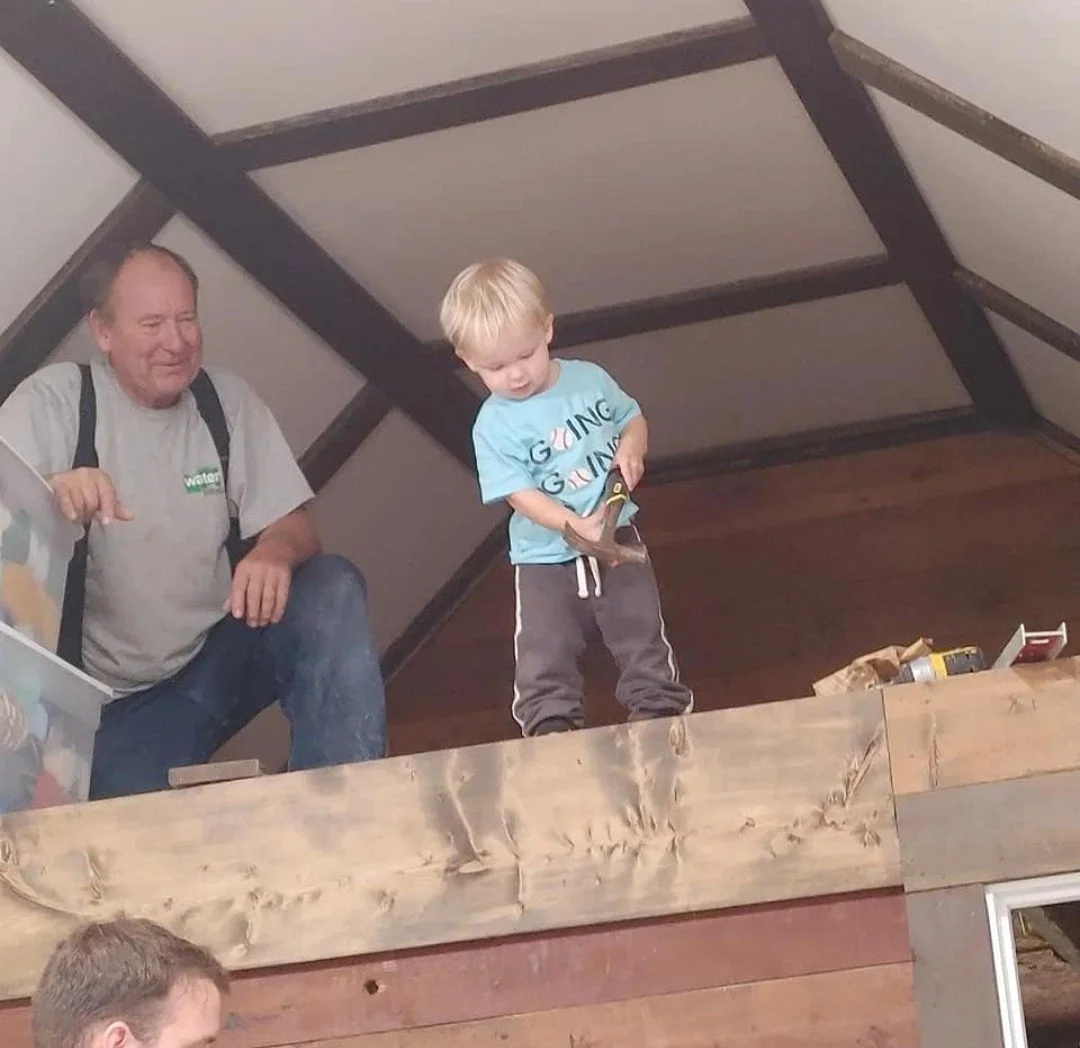 A young boy looking at something in his hands while an older man sits nearby in a room with sloped ceilings and wooden panels.