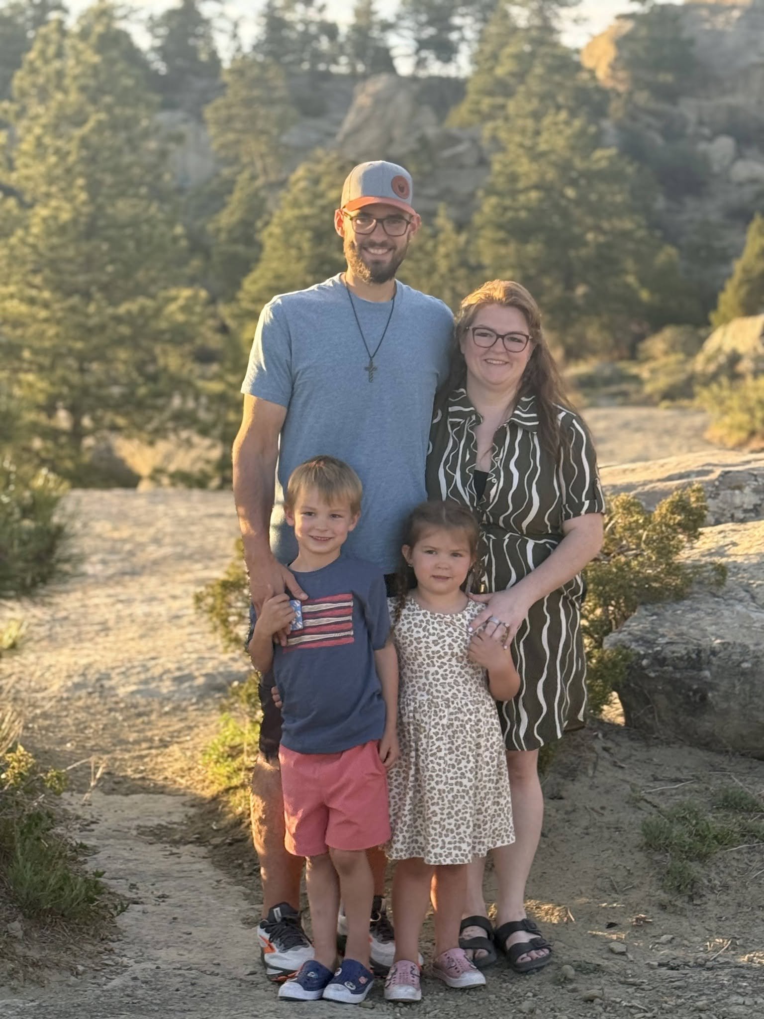 A family of six standing together outdoors on a dirt path, with trees and rocky hills in the background at sunset.
