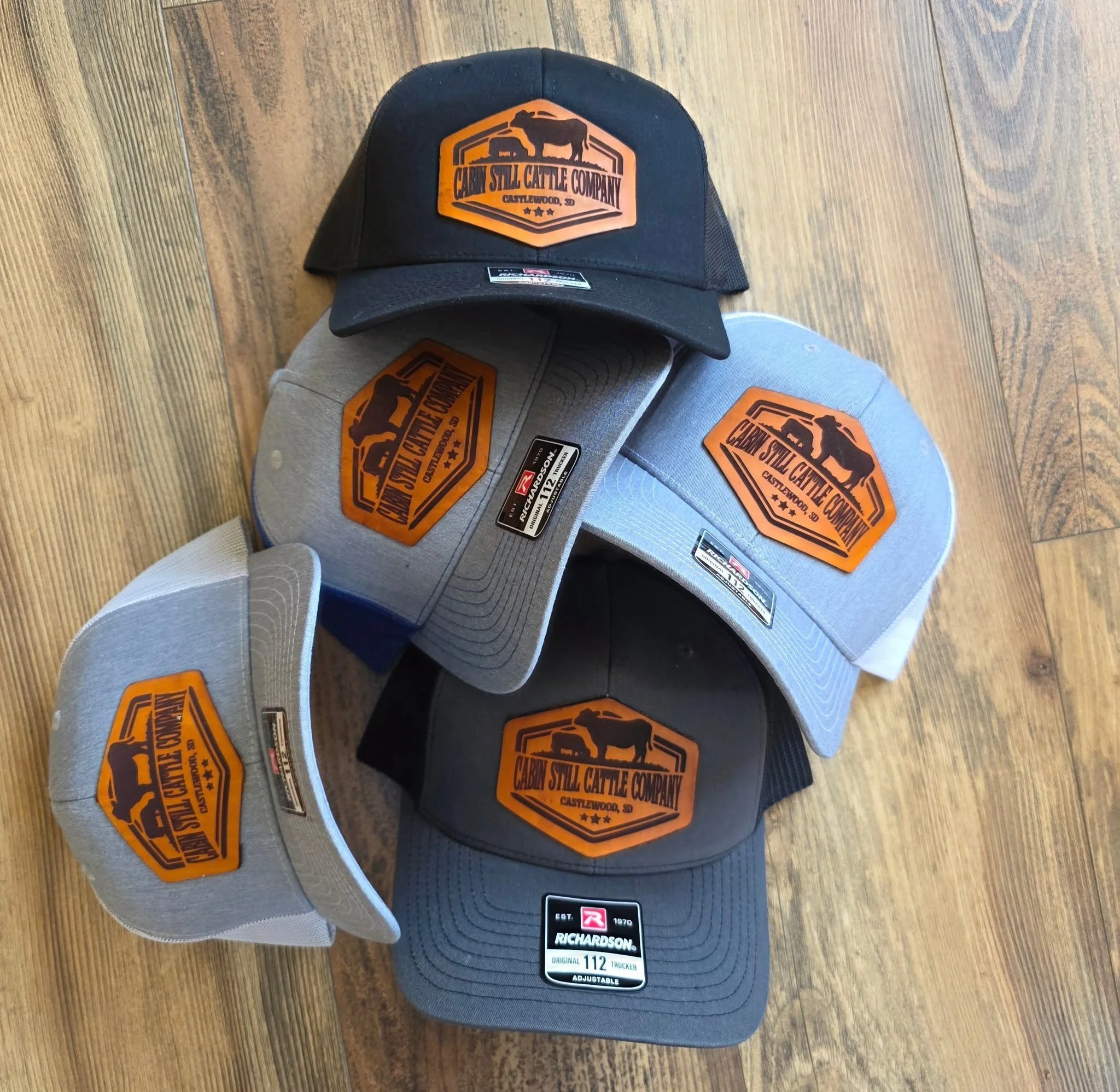 Multiple trucker caps on a wooden floor, featuring patches with the text 'Cattle Still Cattle Company' and a logo of a cow, with various brand and size tags.