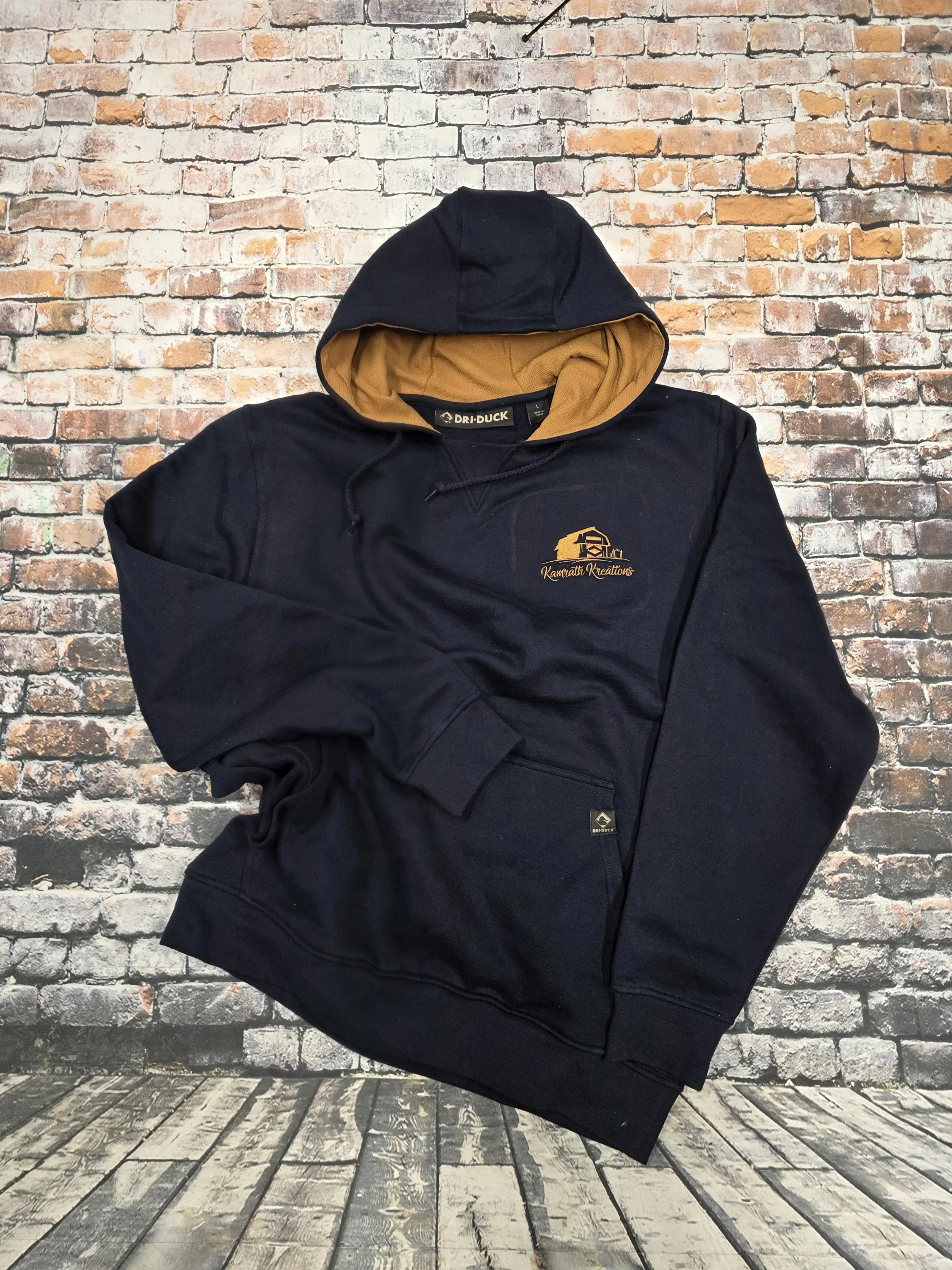 A black hoodie with a tan interior lining, displaying a logo on the left chest area that reads 'Kawatti Kreations' alongside a graphic of a coffee cup, posed against a brick wall background.
