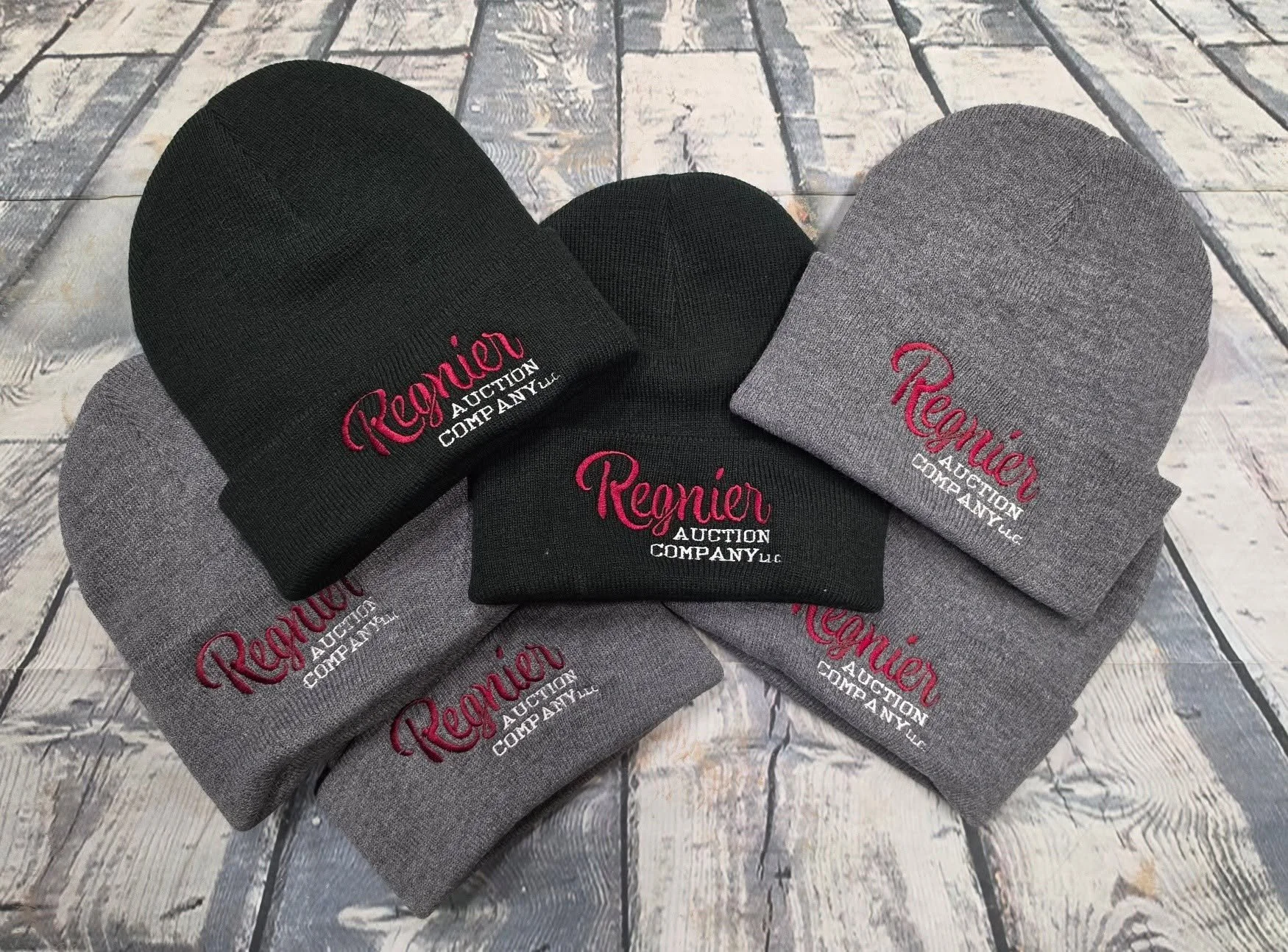 Six knit beanies scattered on a wooden floor, with three in black and three in gray. Each beanie has red and white embroidered text that reads "Reigneur Auction Company LLC."