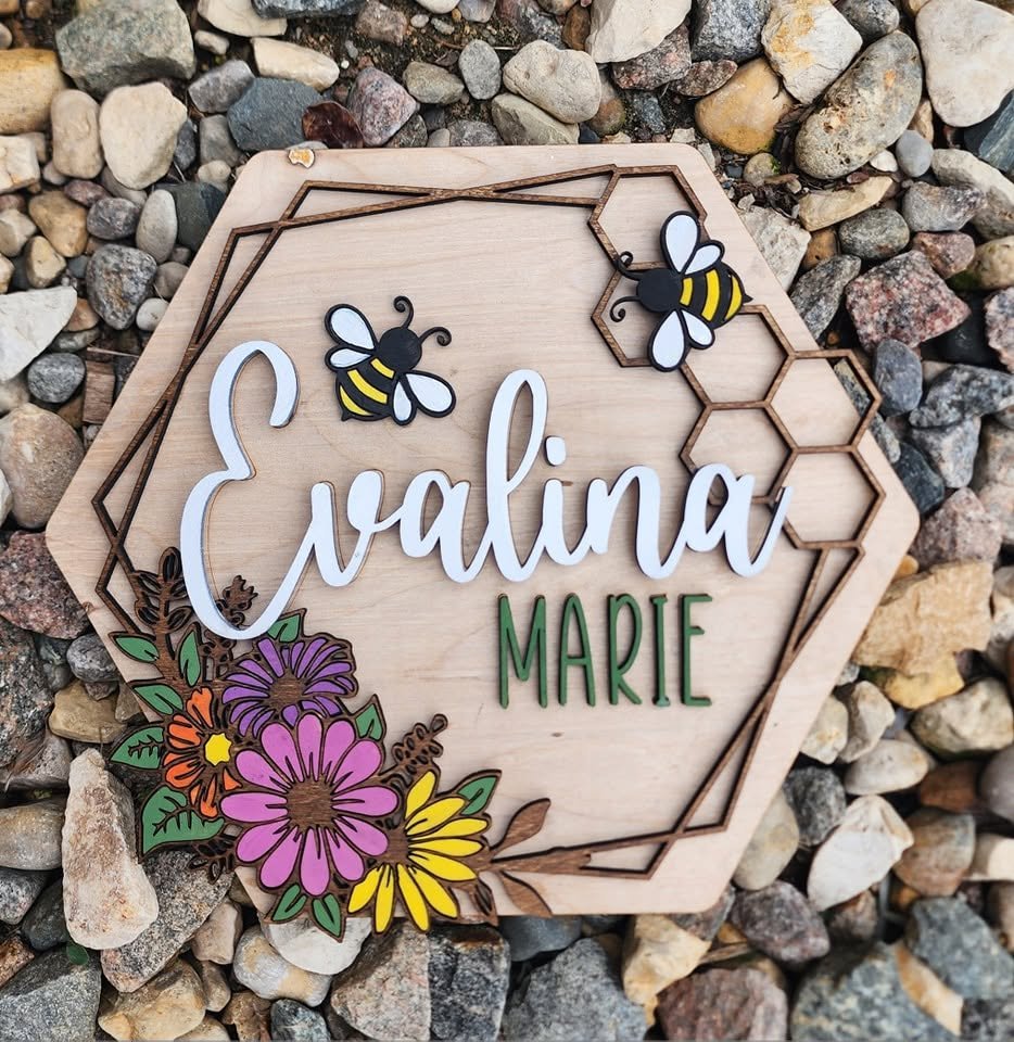 Decorative sign with the name Eva Lina Marie, featuring bees, honeycomb patterns, and colorful flowers, placed on a bed of rocks.