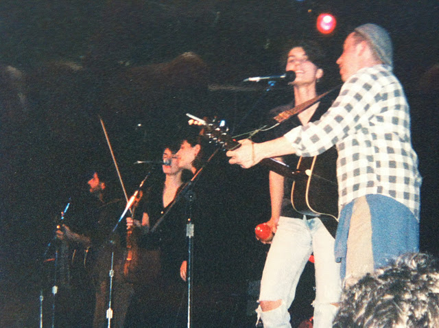 Musicians performing on stage with guitars and microphones, including a woman singing and a man playing guitar in front of an audience.