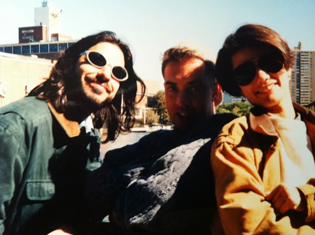 Group of three friends outdoors on a sunny day, wearing sunglasses, smiling, with buildings and blue sky in the background.