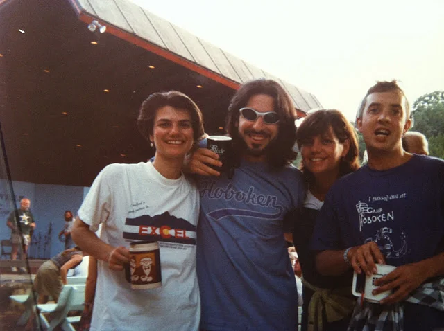 Four smiling friends standing together outdoors during daytime, holding drinks, with other people and a building in the background.