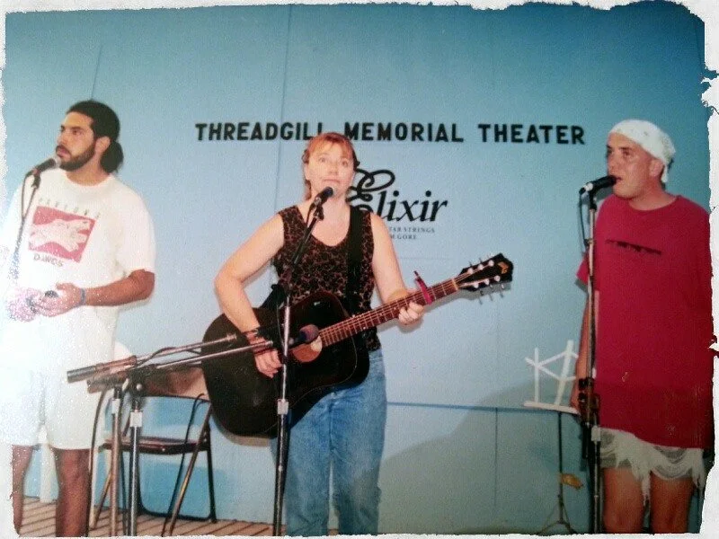 Three people on stage performing at Threadgill Memorial Theater, with one woman playing guitar and singing, and two men standing beside her, one holding a microphone and the other with a white towel on his head, all dressed casually.