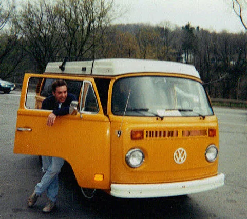 Person leaning out of a yellow Volkswagen van with a white roof, parked outdoors with trees in the background.