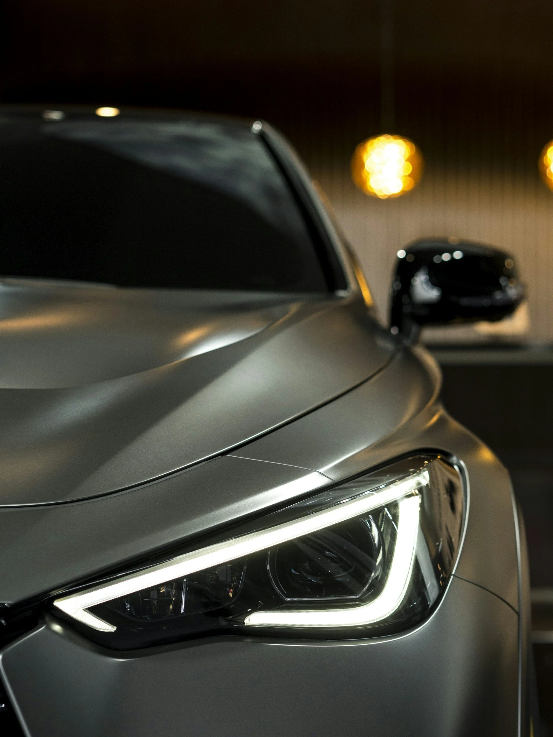 Close-up of a modern silver car's front end, showcasing the headlight and side mirror in a dimly lit environment with warm, out-of-focus lights in the background.