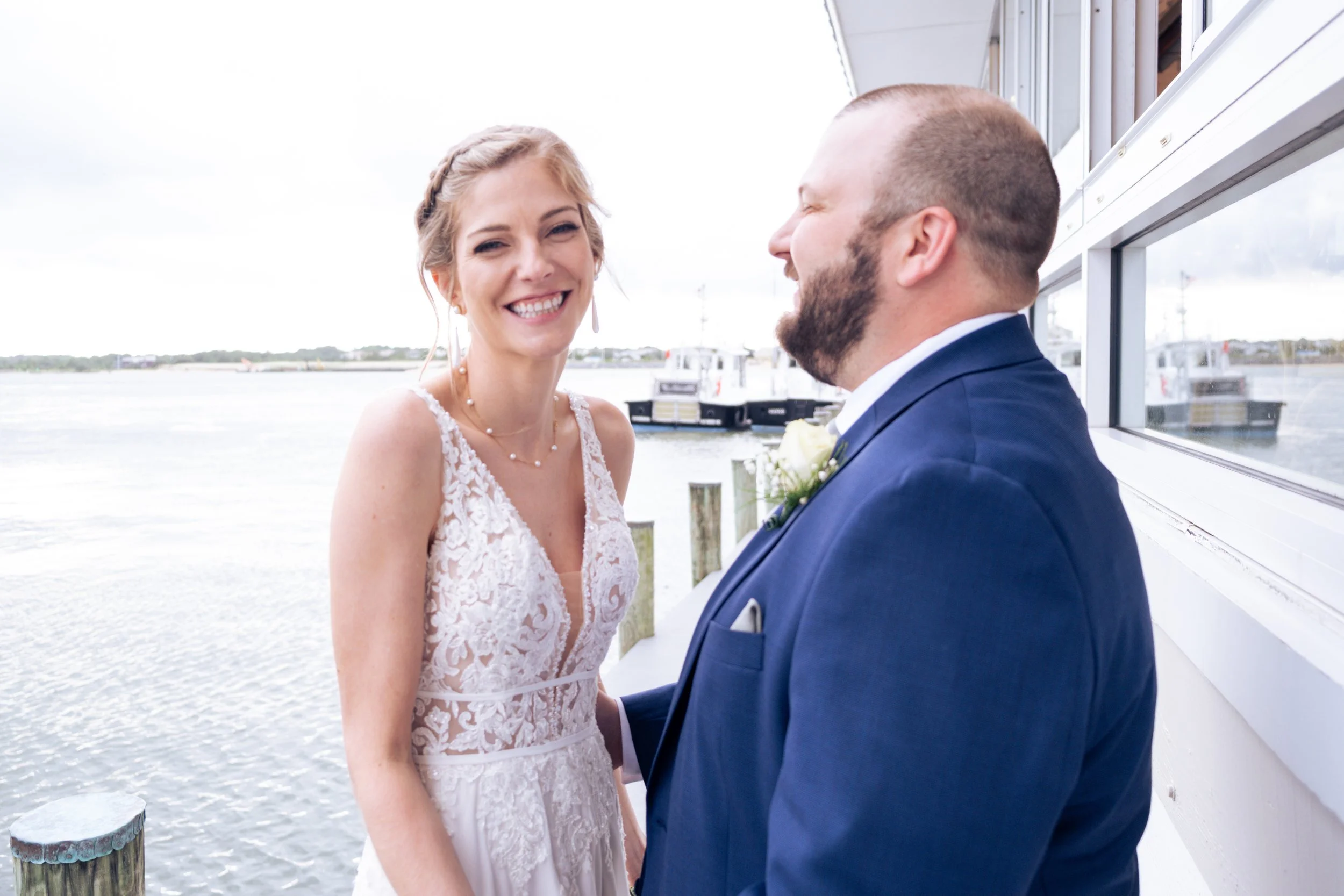 A bride and groom stand facing each other by a waterfront, smiling during a wedding ceremony.