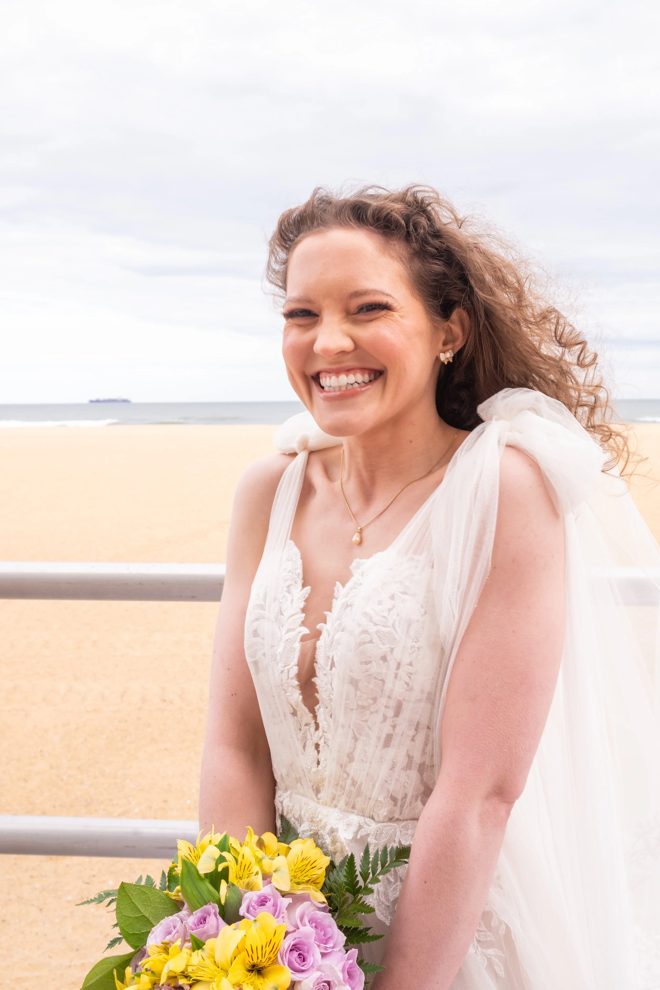 A young woman with curly hair smiling, wearing a white dress, holding a bouquet of yellow and purple flowers, on a beach with cloudy sky and distant ship.