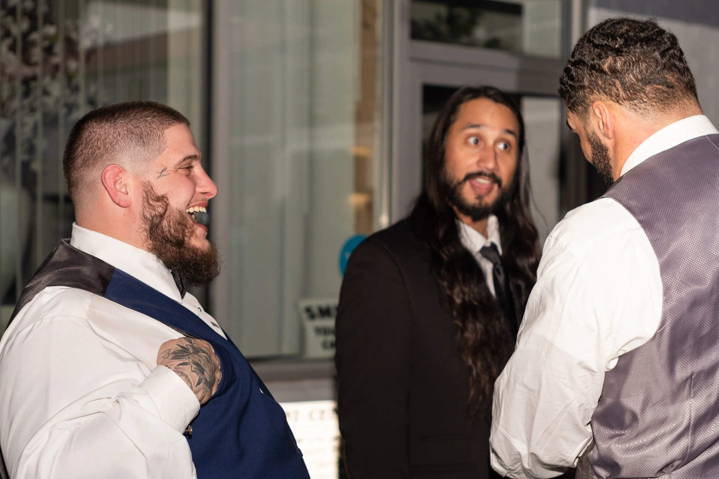 Three men dressed in formal and business casual attire are engaged in a lively conversation, smiling and laughing indoors.