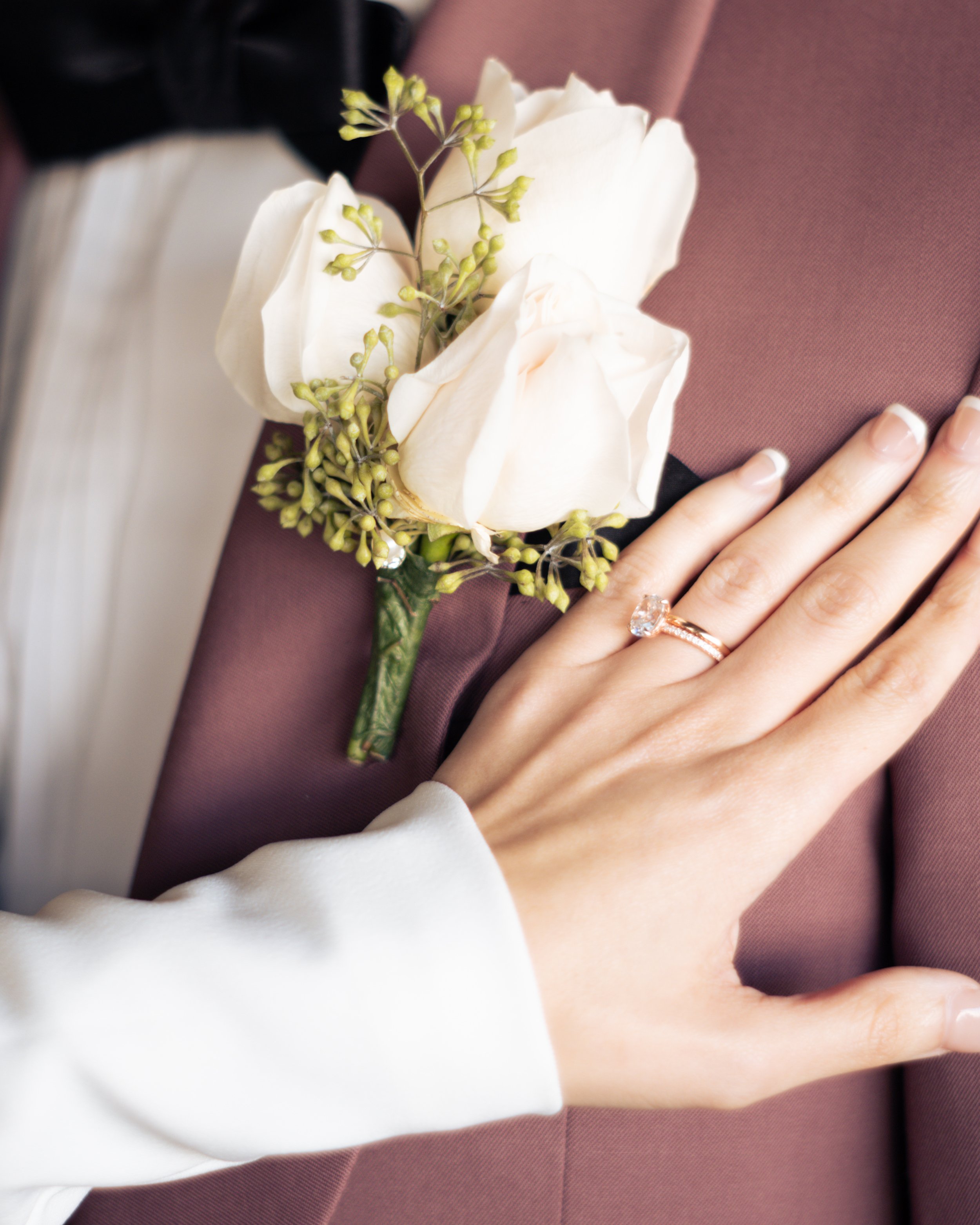 Close-up of a person's hand with a wedding ring resting on a brown fabric, with a white flower and green foliage nearby.