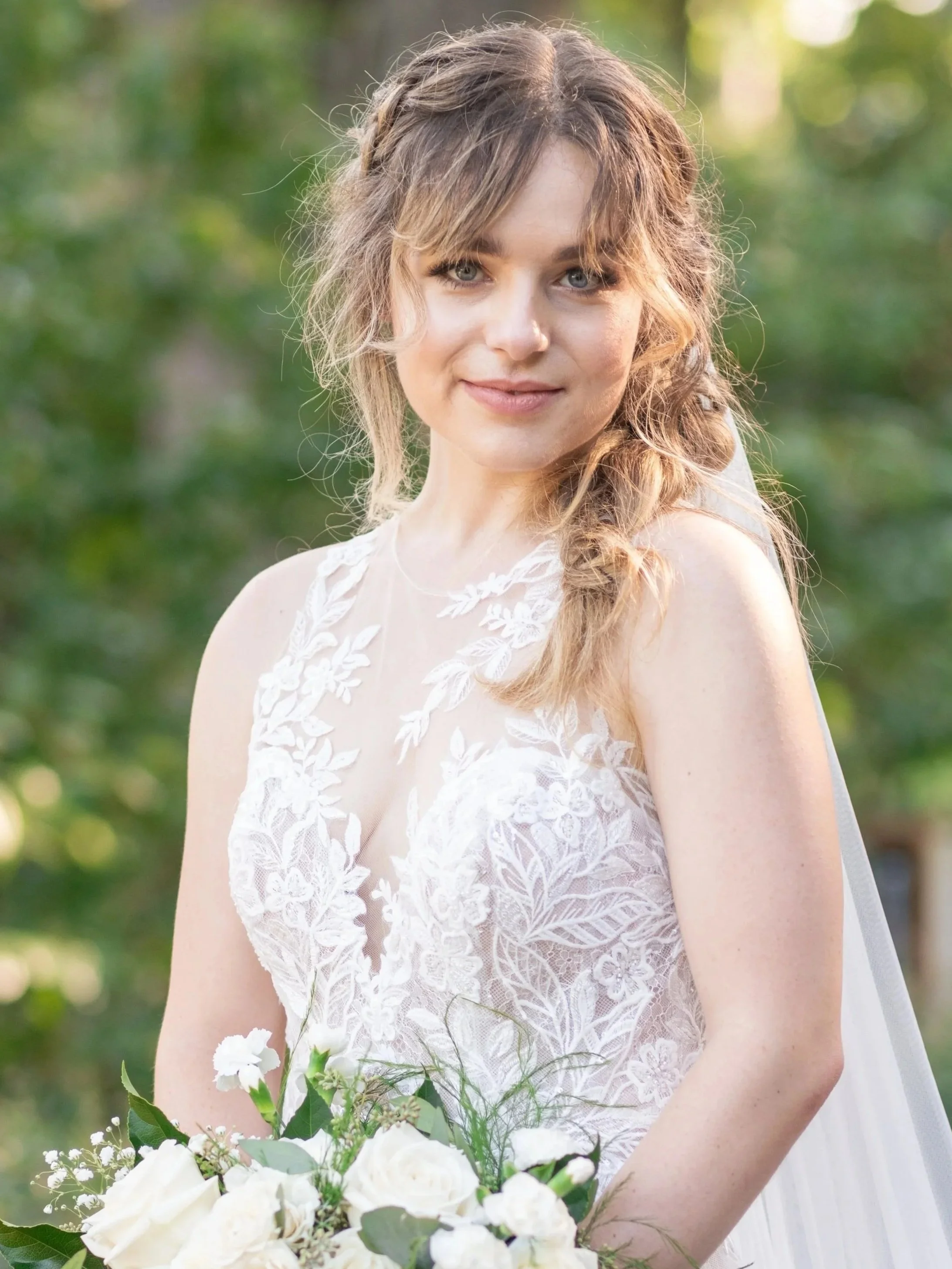 A bride with wavy hair in a loose braid, wearing a white lace wedding dress, holding a bouquet of white flowers with greenery, standing outdoors in a green, blurred background.