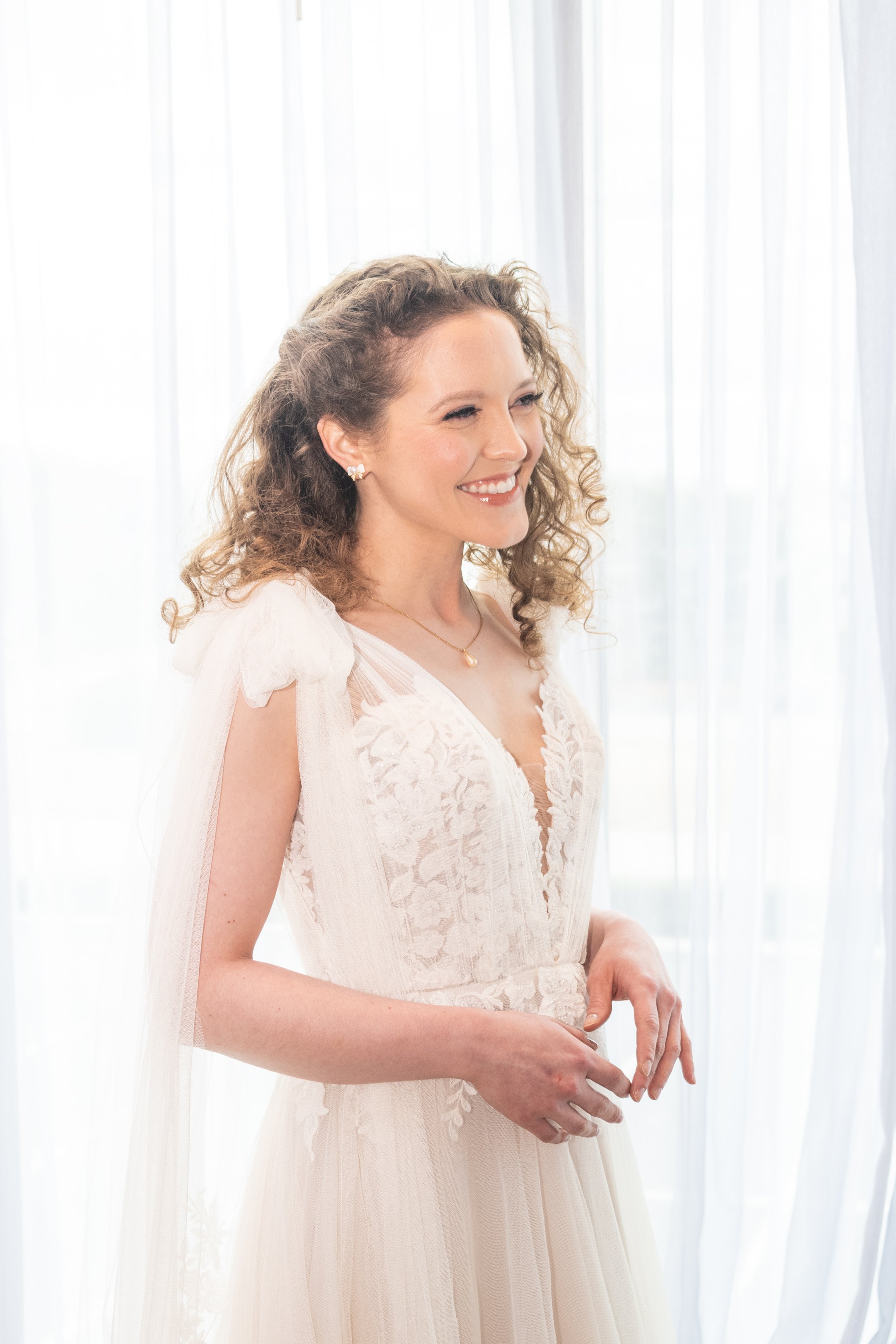 A woman in a white wedding dress with lace detailing, standing by white curtains, smiling gently with her hands clasped in front of her.