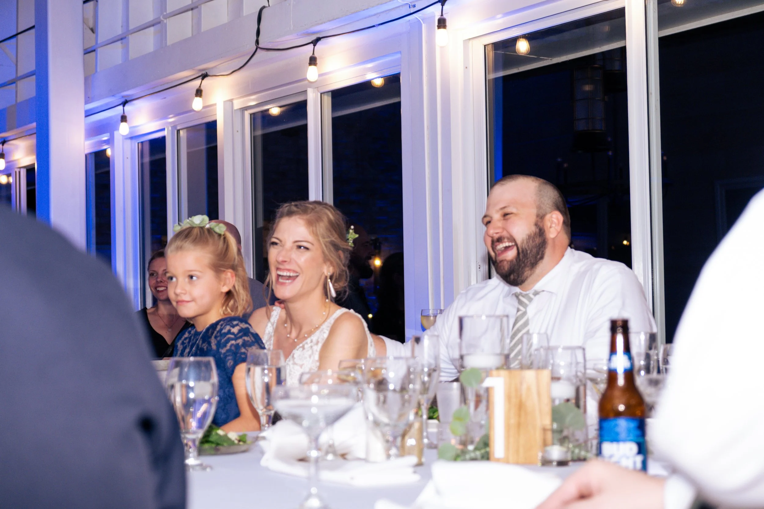 People dressed in formal attire sitting at a long table with wine glasses, bottles, and decorations, enjoying a gathering or celebration indoors illuminated by hanging string lights.