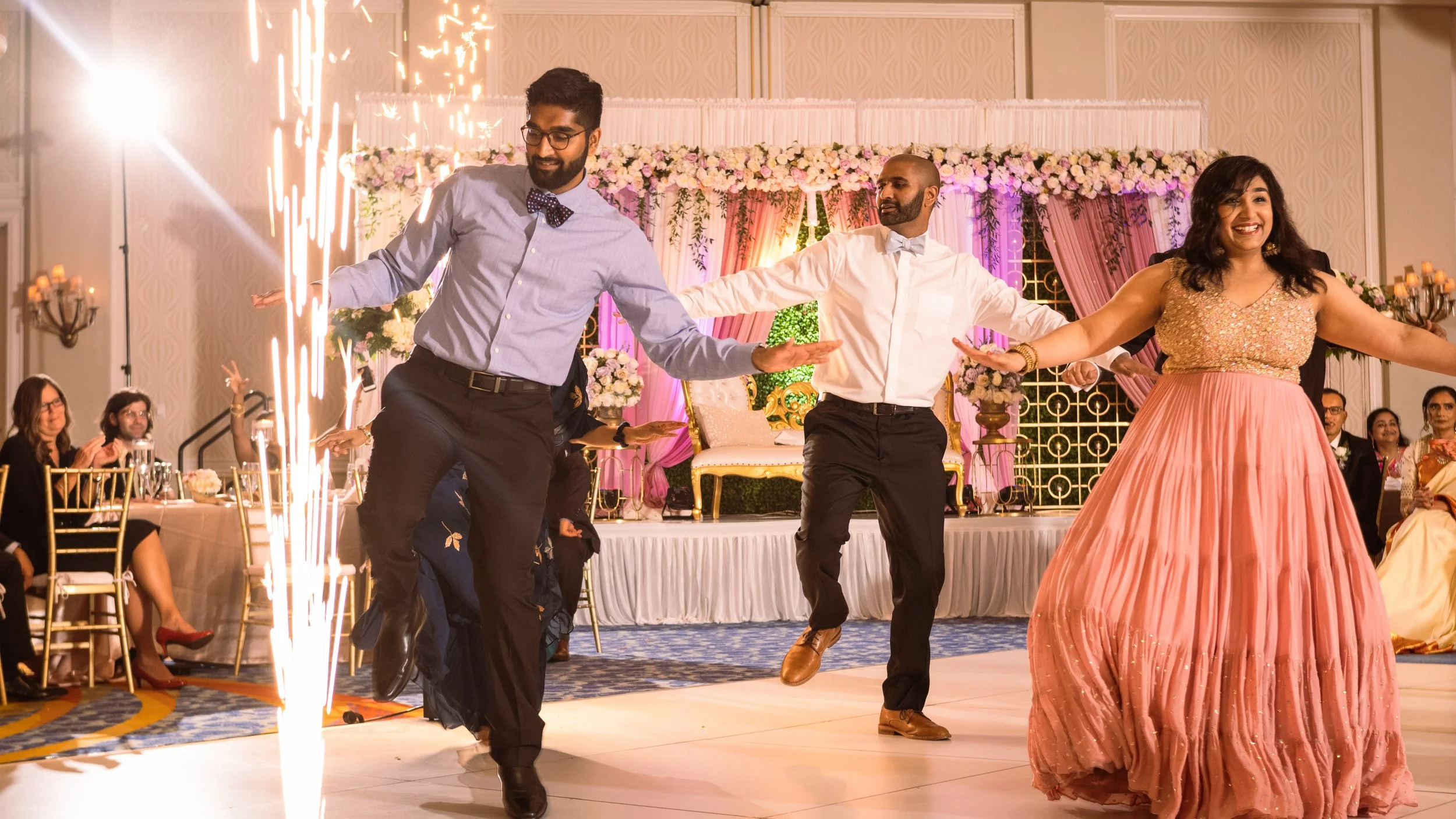 People dancing at a wedding reception, with a decorated stage and seated guests in the background.