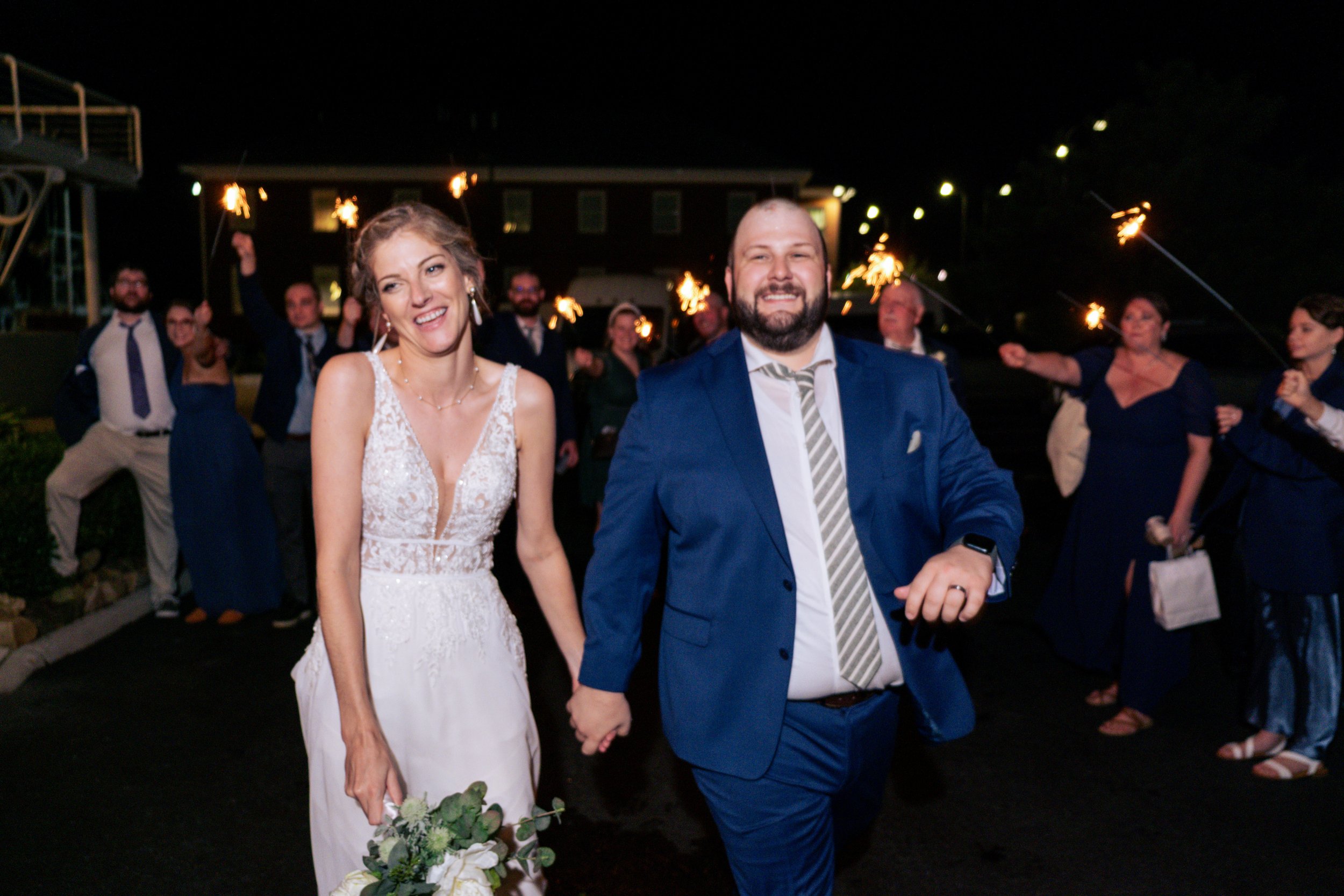 A newly married couple holding hands at night surrounded by friends and family holding sparklers, celebrating outdoors.