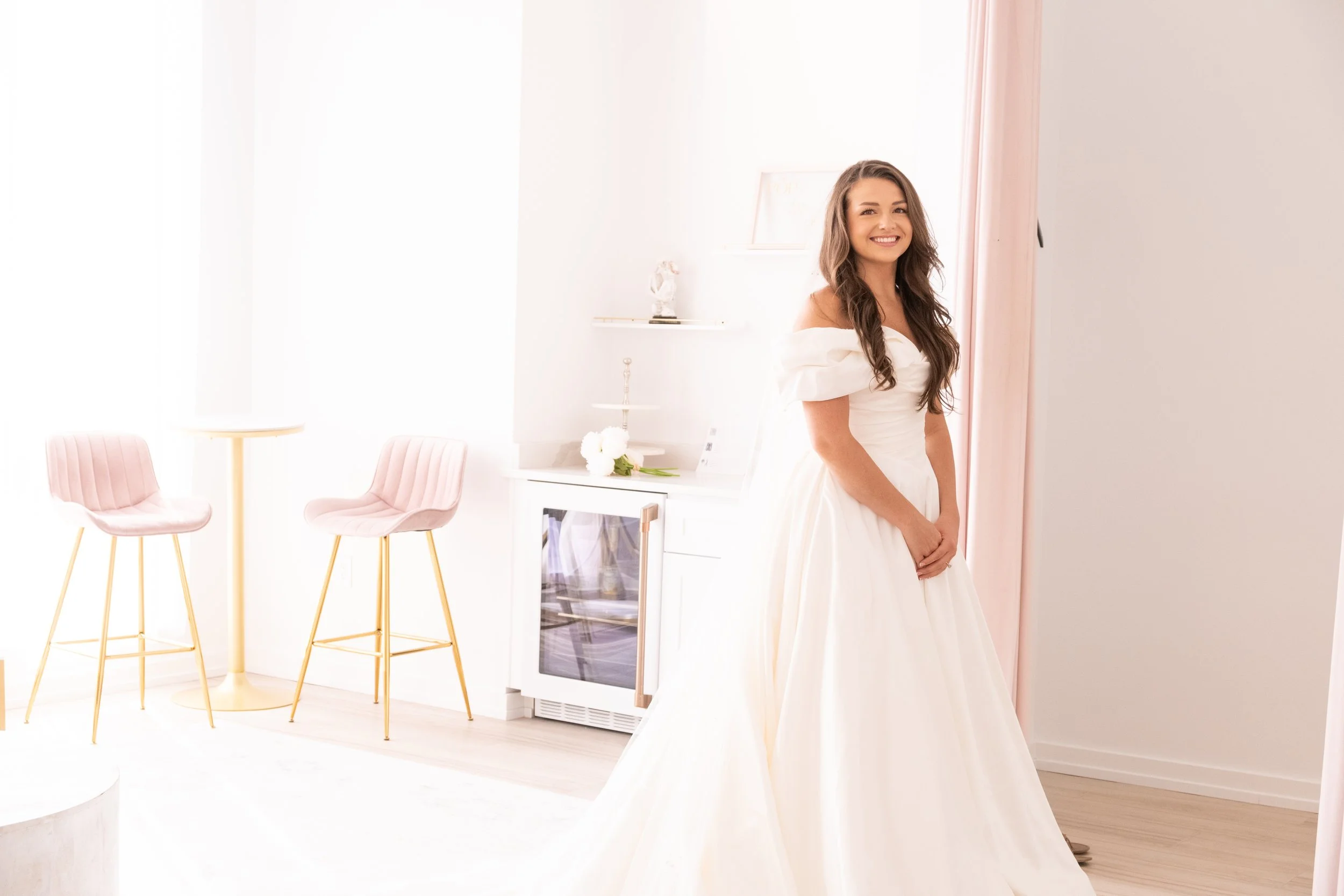 A woman in a white off-shoulder dress smiling in a bright, modern room with pink chairs and white decor.