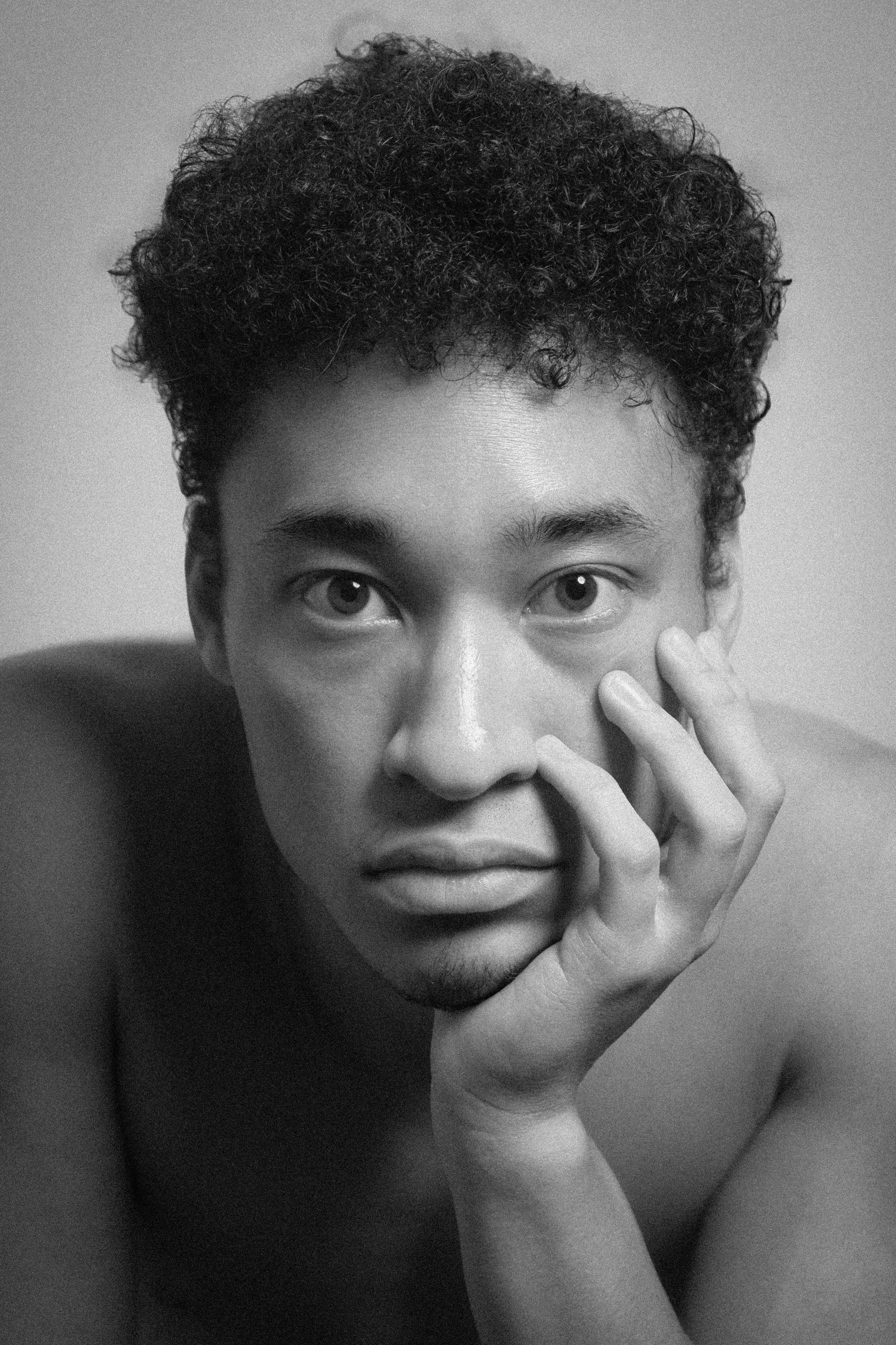 Black and white portrait of a young person with short curly hair, looking directly at the camera, with one hand touching their face.