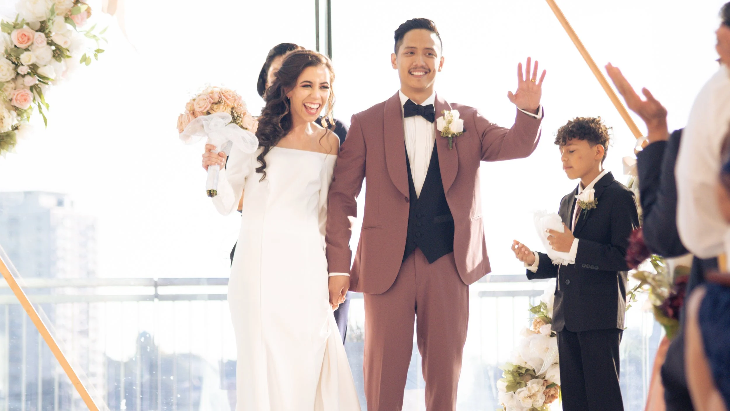 A wedding ceremony with a bride and groom holding hands and smiling as they face the officiant, surrounded by guests and floral decorations.
