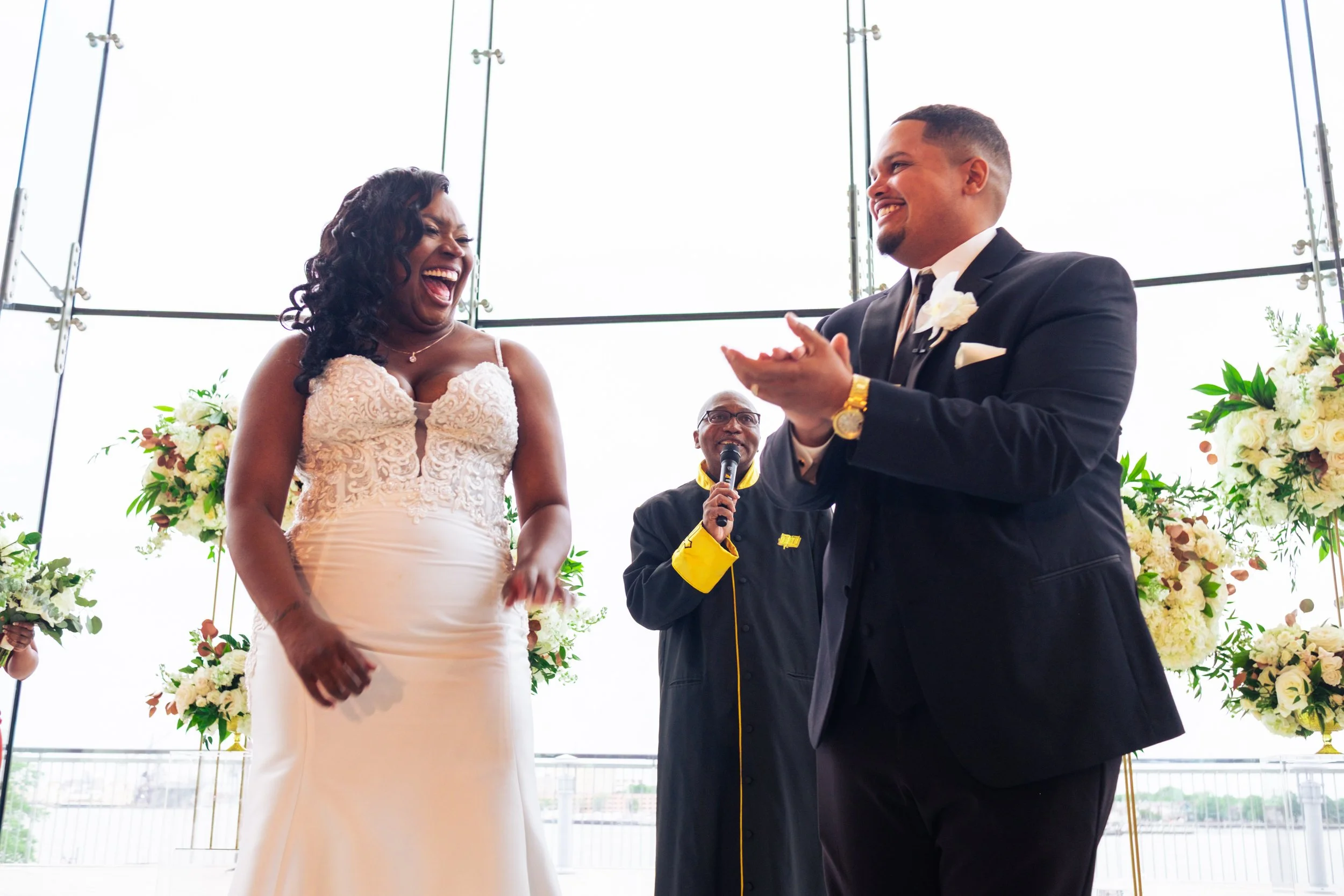 A bride and groom are smiling and clapping during their wedding ceremony inside a glass-walled venue, decorated with floral arrangements.
