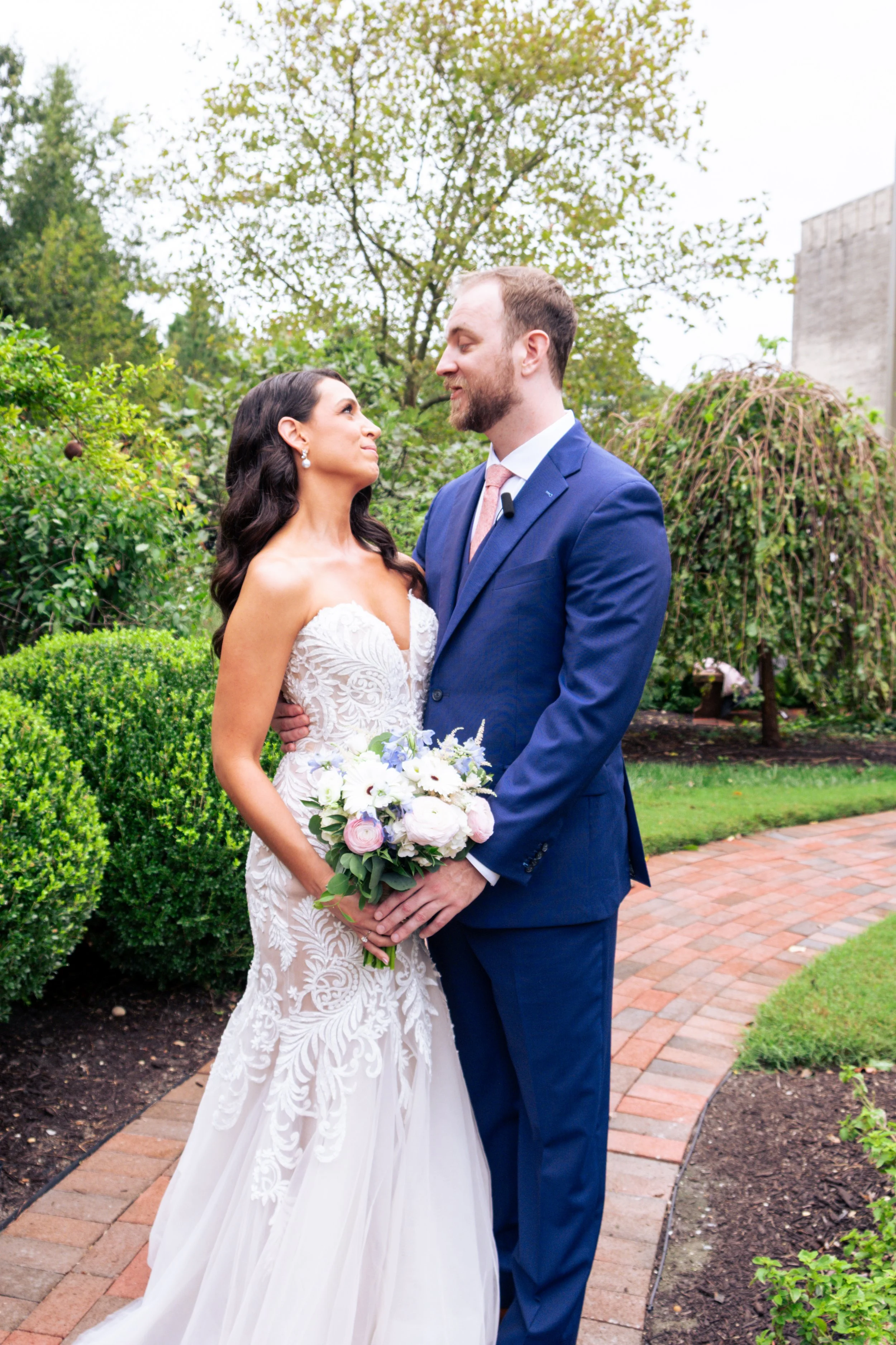 A bride and groom standing together outdoors, gazing into each other's eyes. The bride is wearing a white wedding dress and holding a bouquet of white and pink flowers. The groom is dressed in a blue suit and pink tie. They are surrounded by green sh