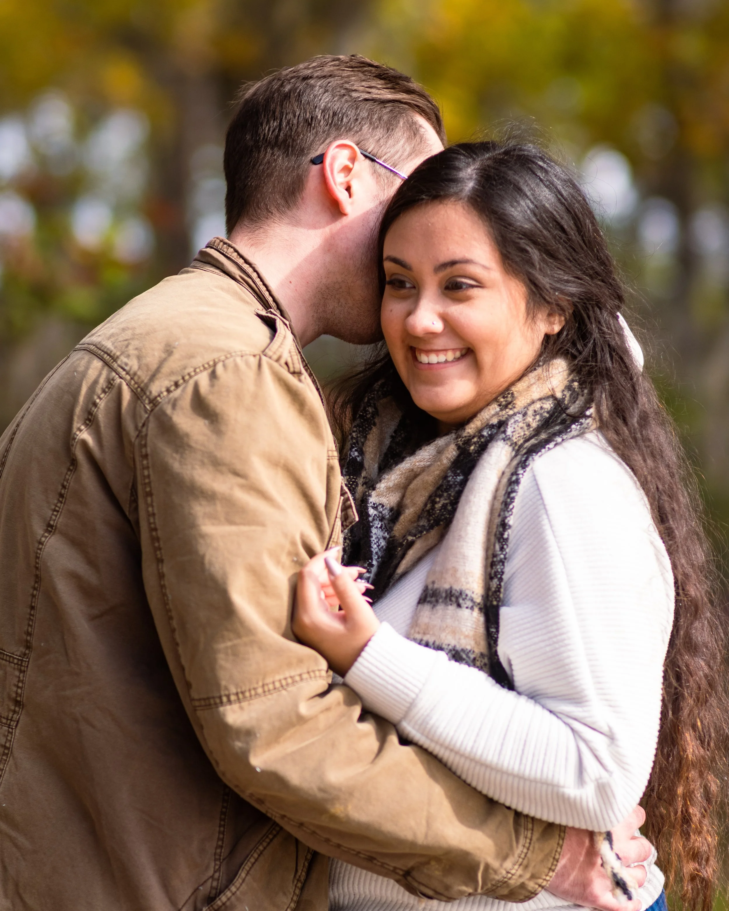 A man with short brown hair and glasses is kissing a smiling woman with dark hair and a nose piercing. They are outdoors with a blurred autumn background.