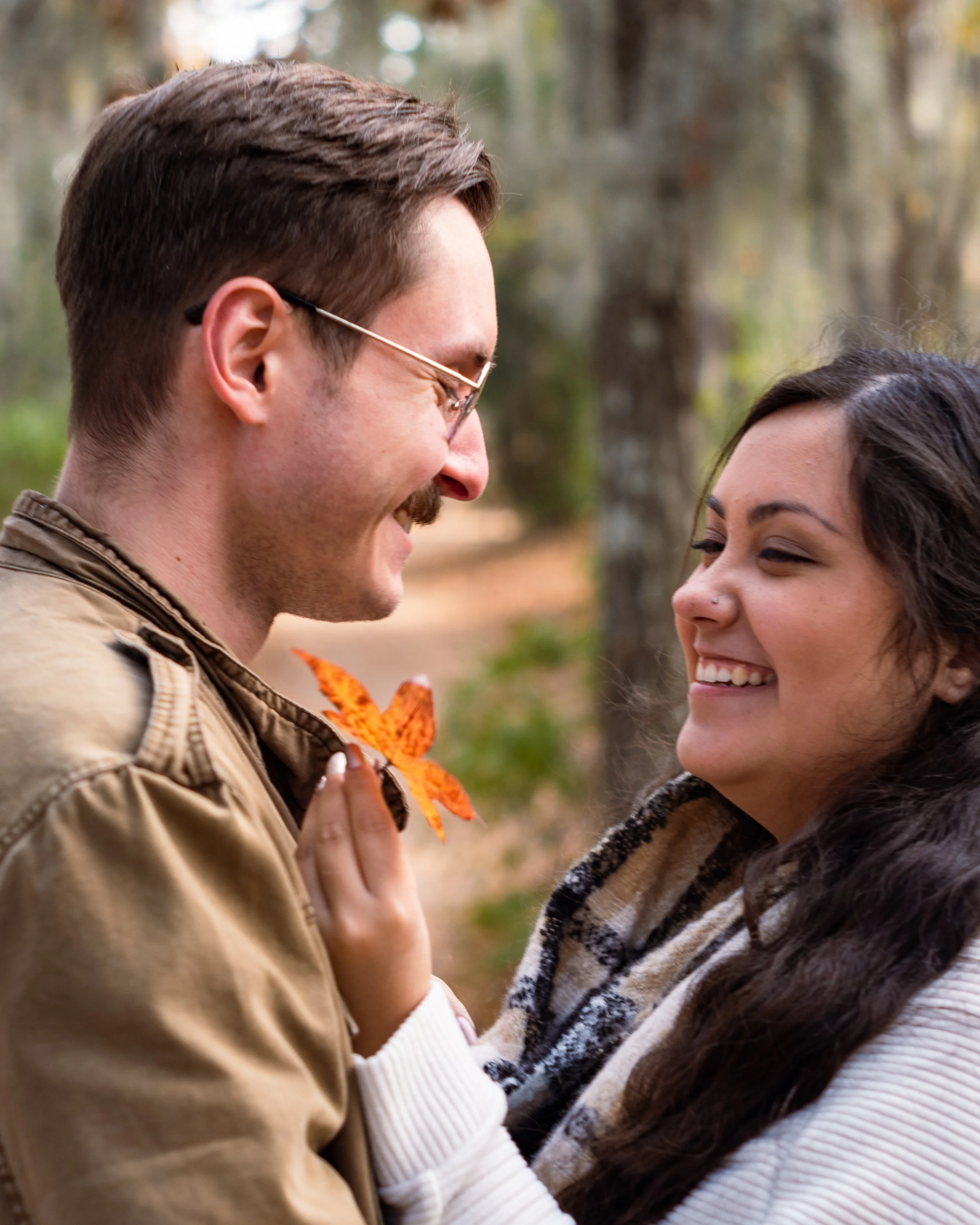 A man and woman face each other outdoors, smiling and close, with blurred trees in the background.